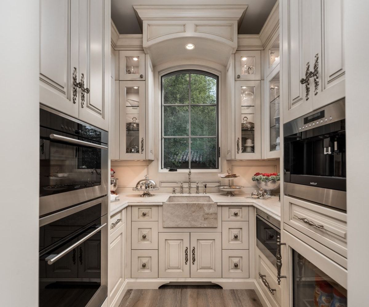 Luxury butler’s pantry with custom cabinets, marble counters, built-in ovens, and arched window