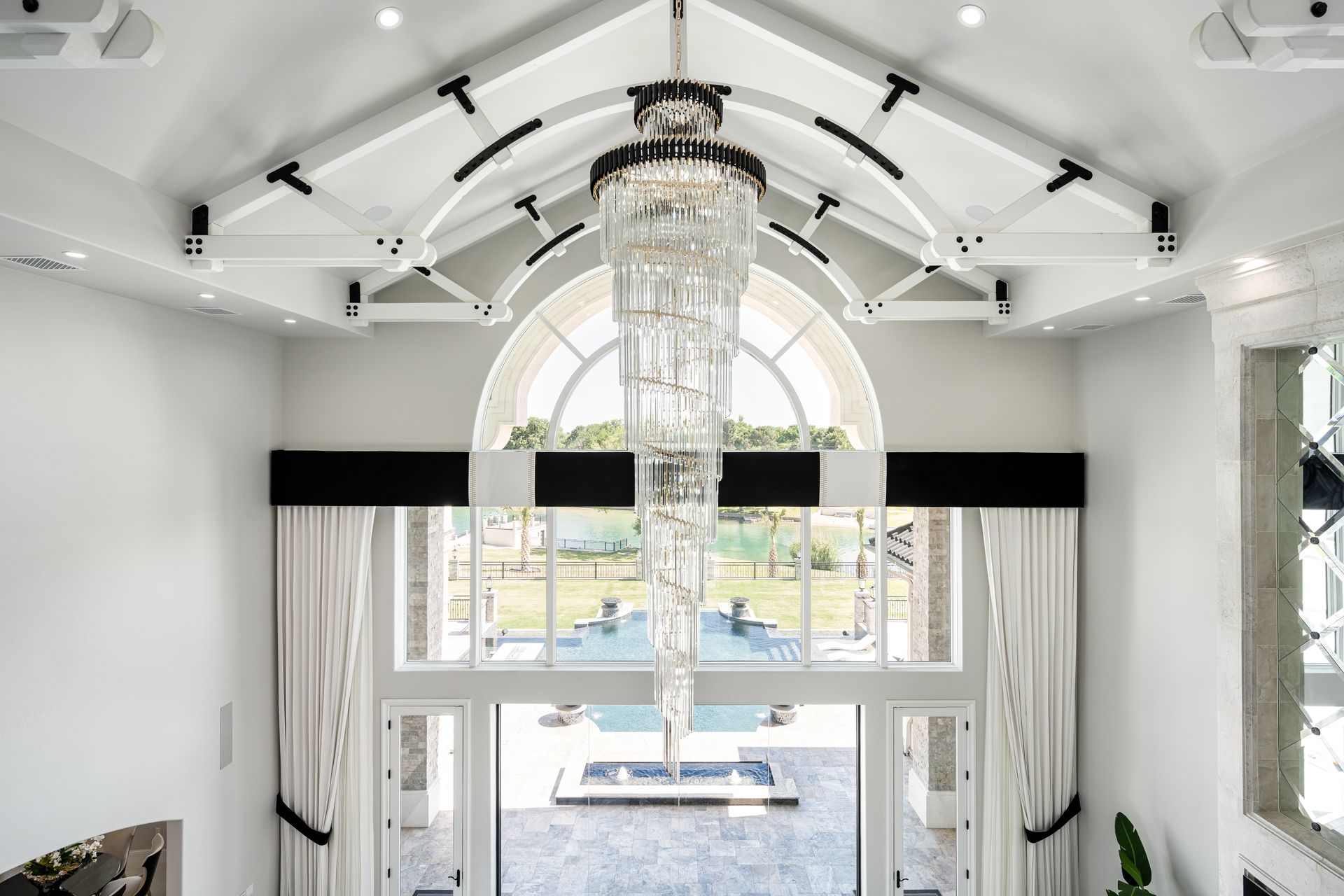 A tall, crystal spiral chandelier hangs beneath a white vaulted ceiling in a room overlooking a swimming pool.