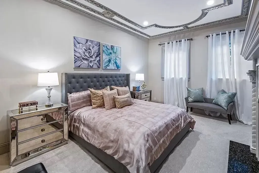 Luxurious bedroom with mirrored furniture, gray tufted headboard, and decorative ceiling trim.