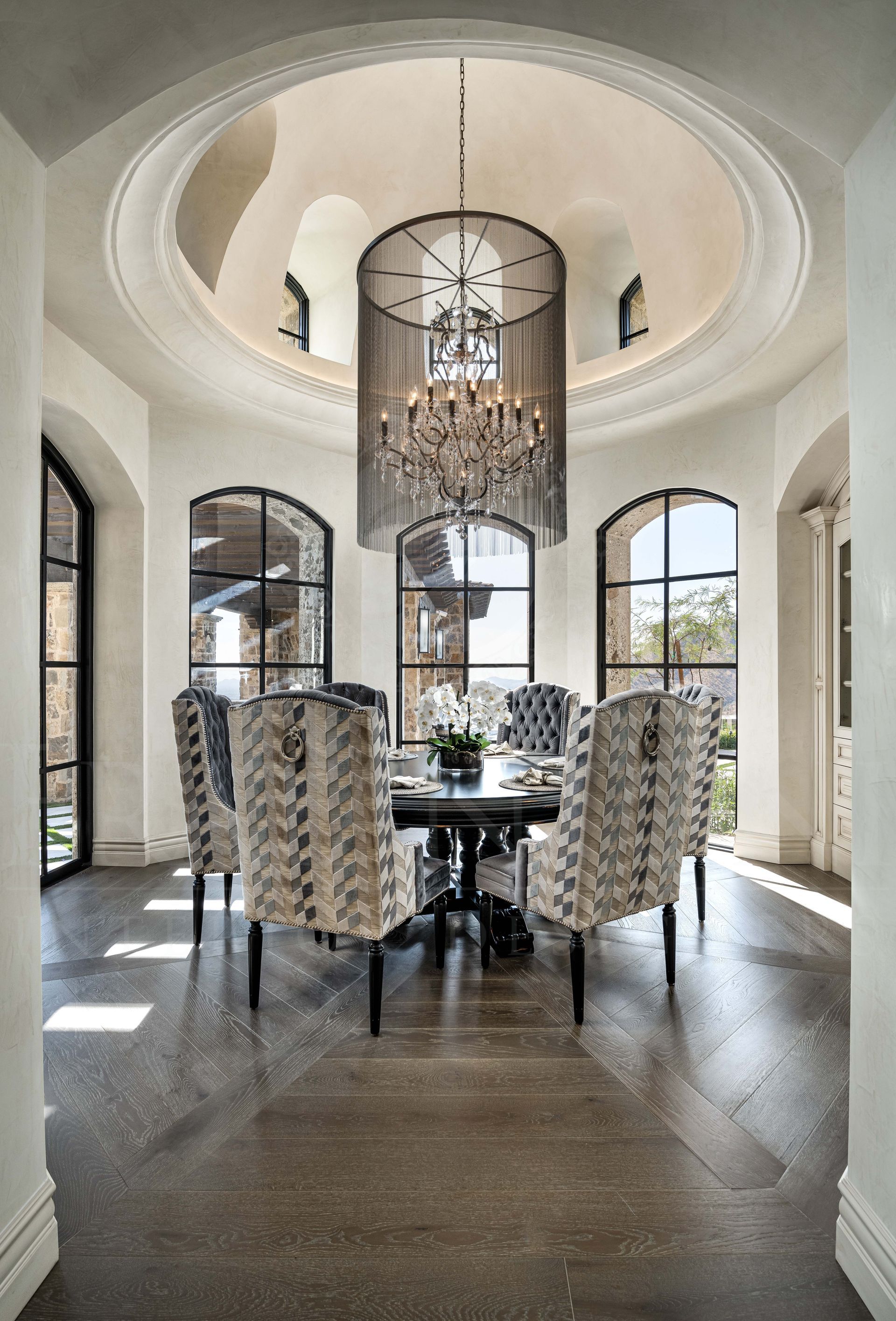 Luxury breakfast nook with chandelier in a vaulted dome ceiling, round table, patterned chairs, arched windows, and wood floors