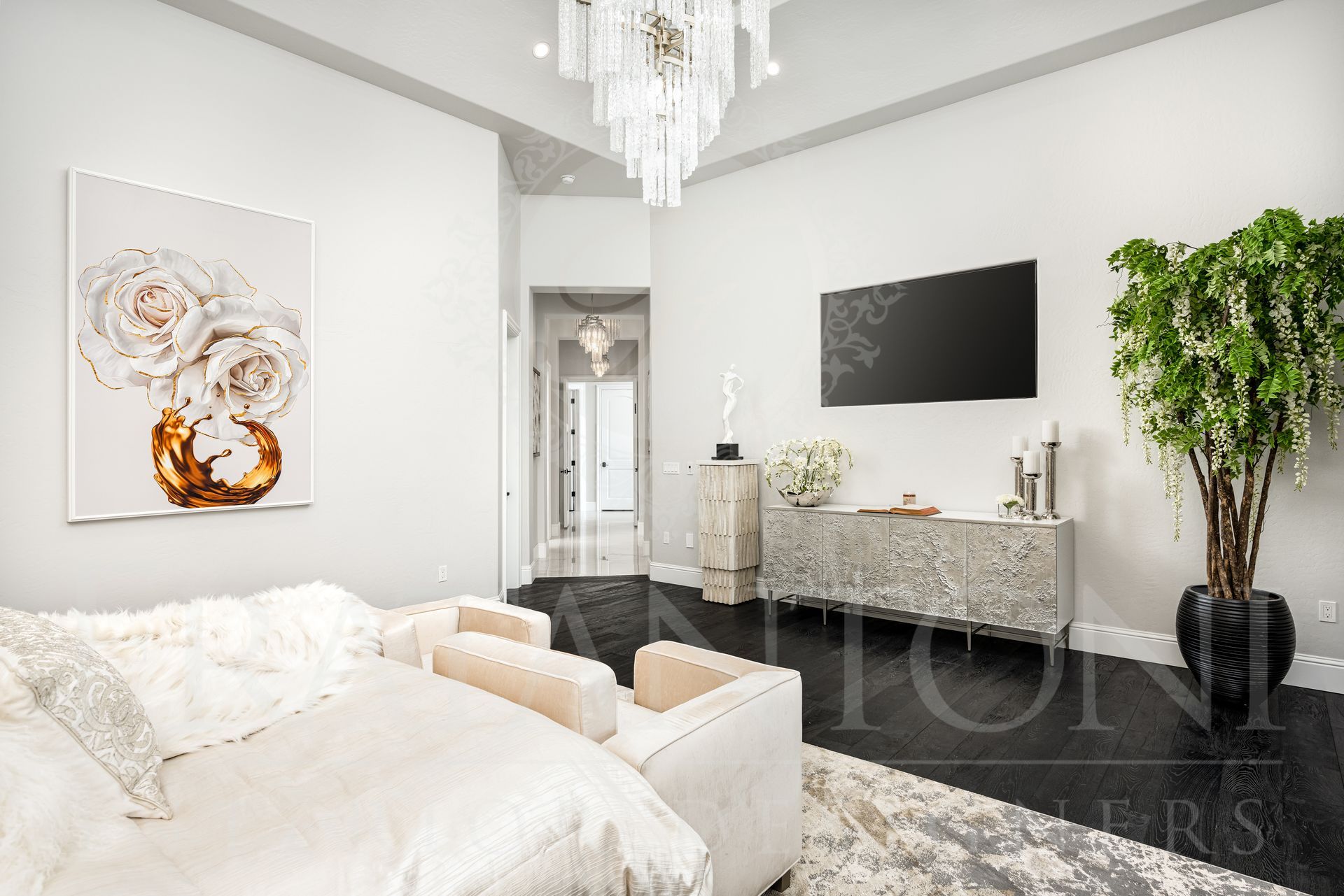 Luxurious bedroom with dark wood flooring, white walls, and ornate chandelier. Features a large TV and a decorative dresser.