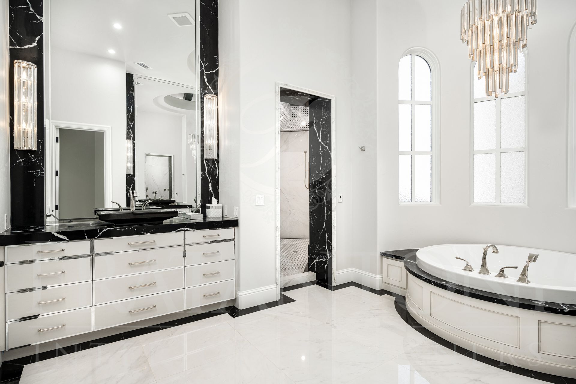 Elegant white and black bathroom with a double vanities, large mirrors, a chandelier, a large walk-in shower, and a bathtub.