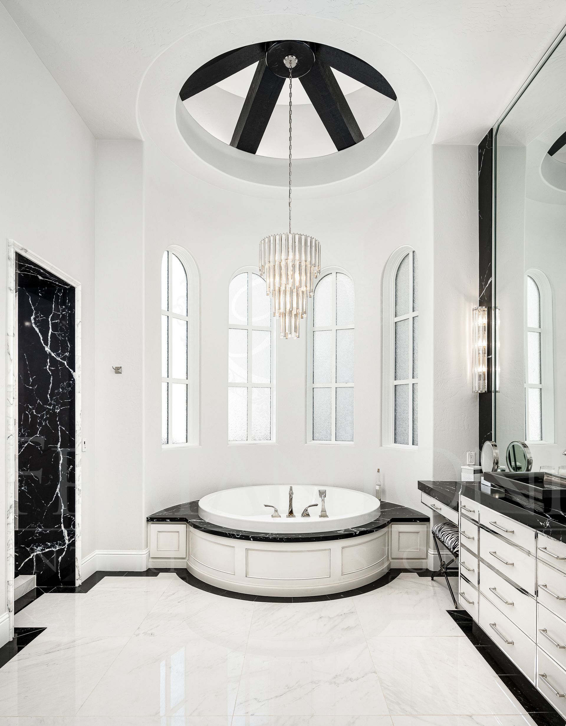 Elegant, white and black bathroom with a round bathtub under arched windows. A chandelier hangs from a ceiling dome designed by Fratantoni Interior Designers.