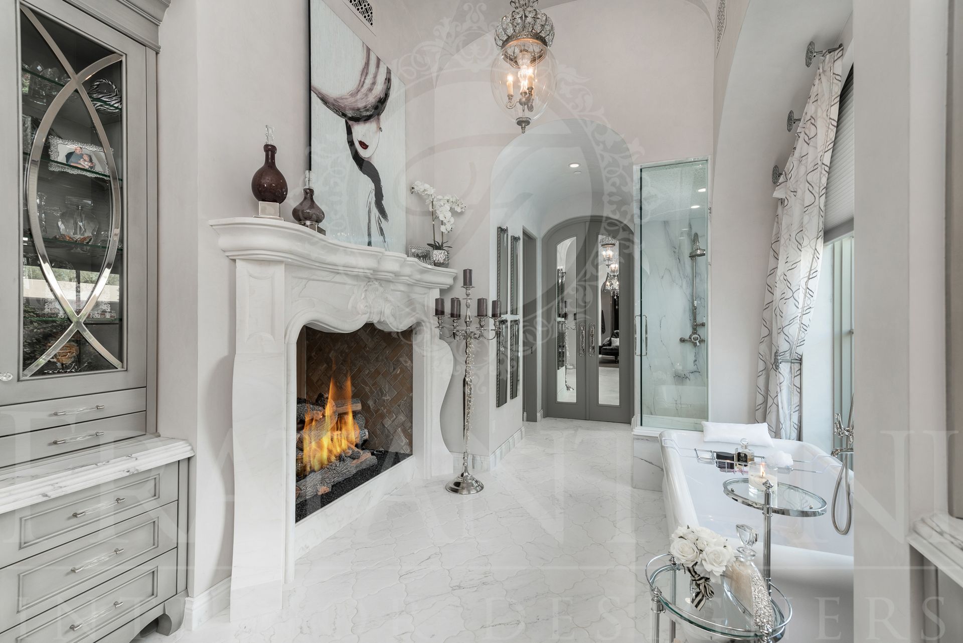 Elegant, all-white bathroom with fireplace, ornate decor, and archways, creating a luxurious and bright ambiance. Also features a freestanding bathtub under a large arched window and a glass shower. Located in SIlverleaf and designed by Fratantoni Interior Designers in Scottsdale, Arizona