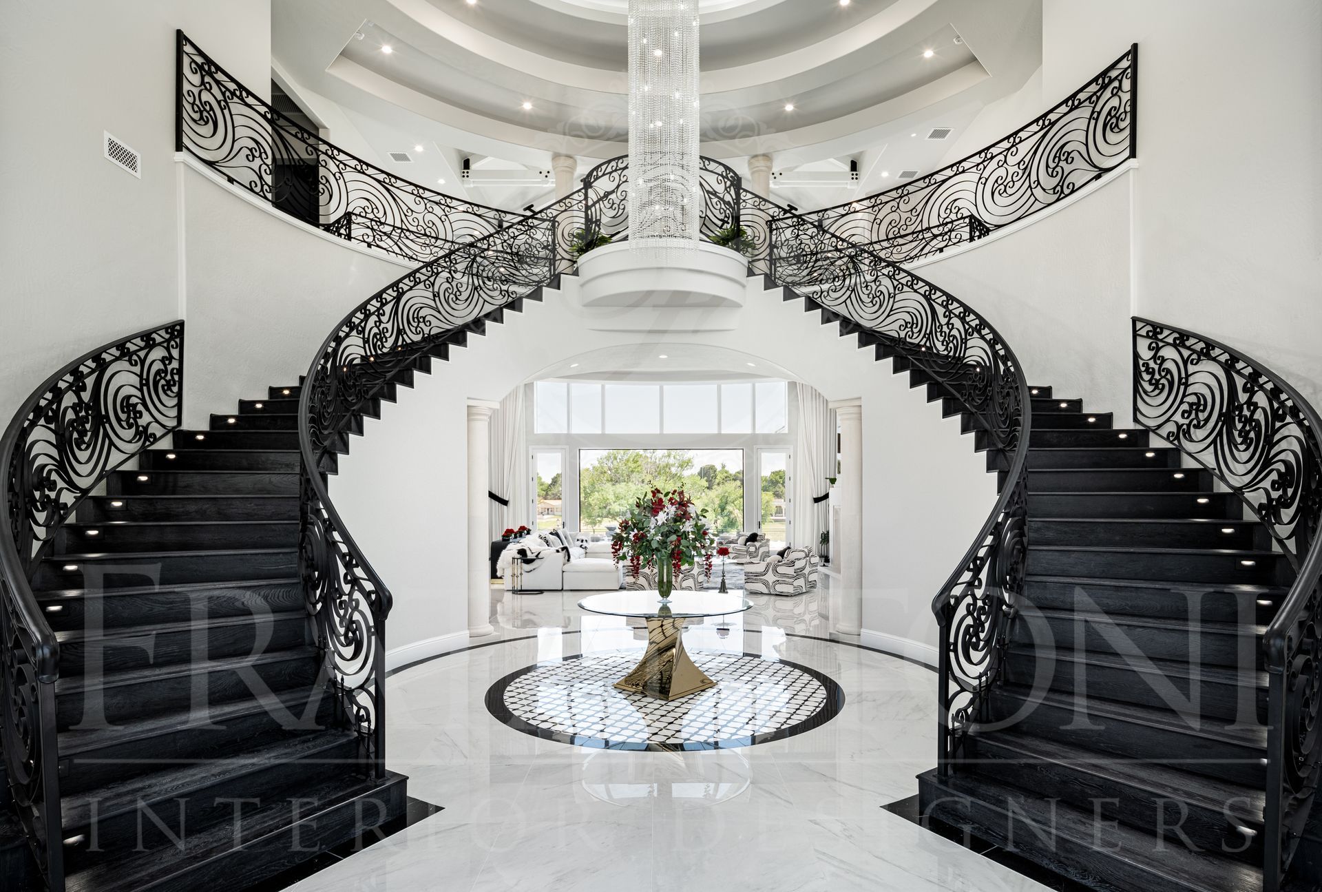 Elegant foyer with dual curved staircases, black ornate railings, chandelier, and a central round floor design.