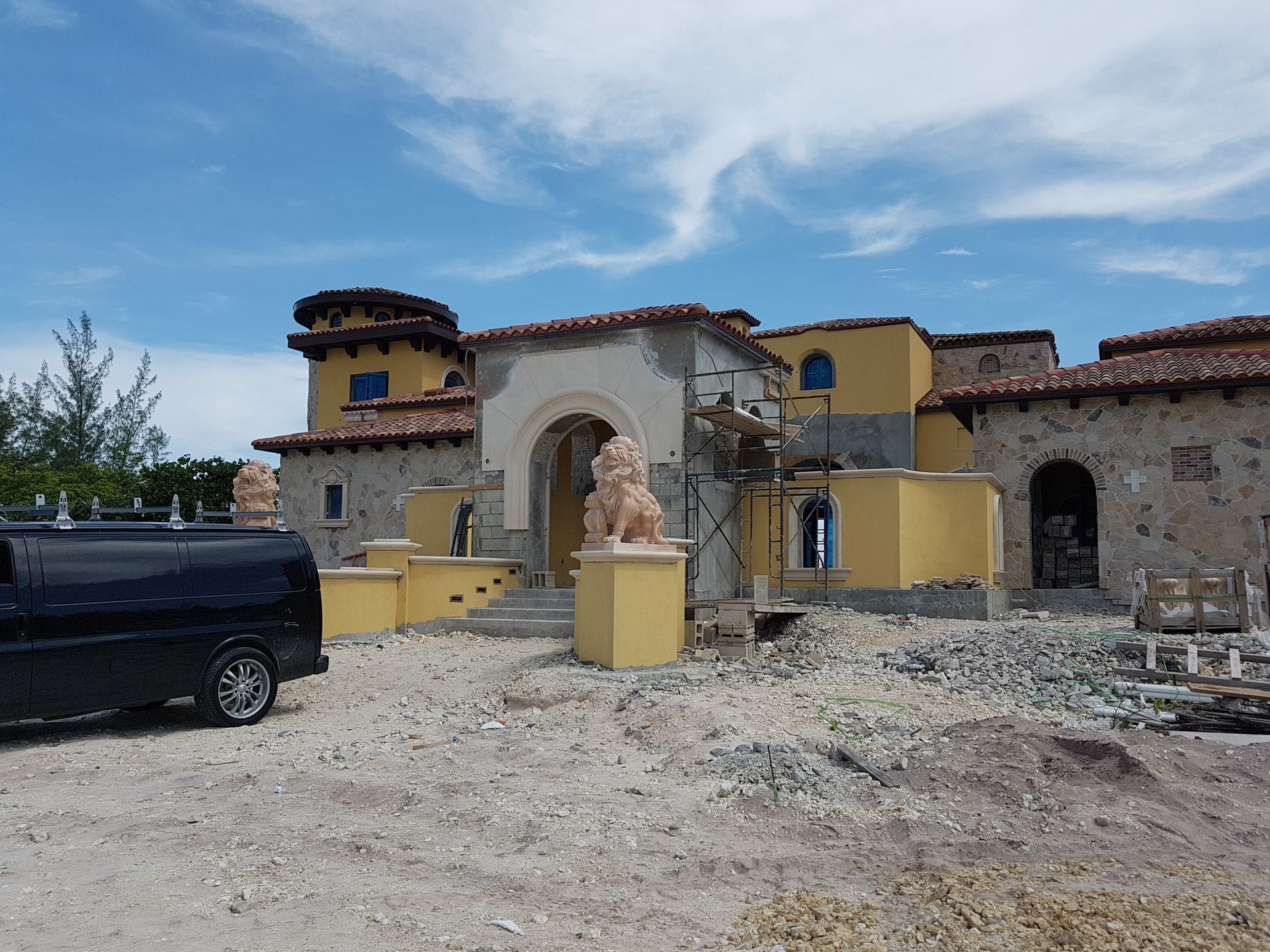 Exterior view of a large, unfinished stucco building with stone accents; a black van is parked in the foreground.