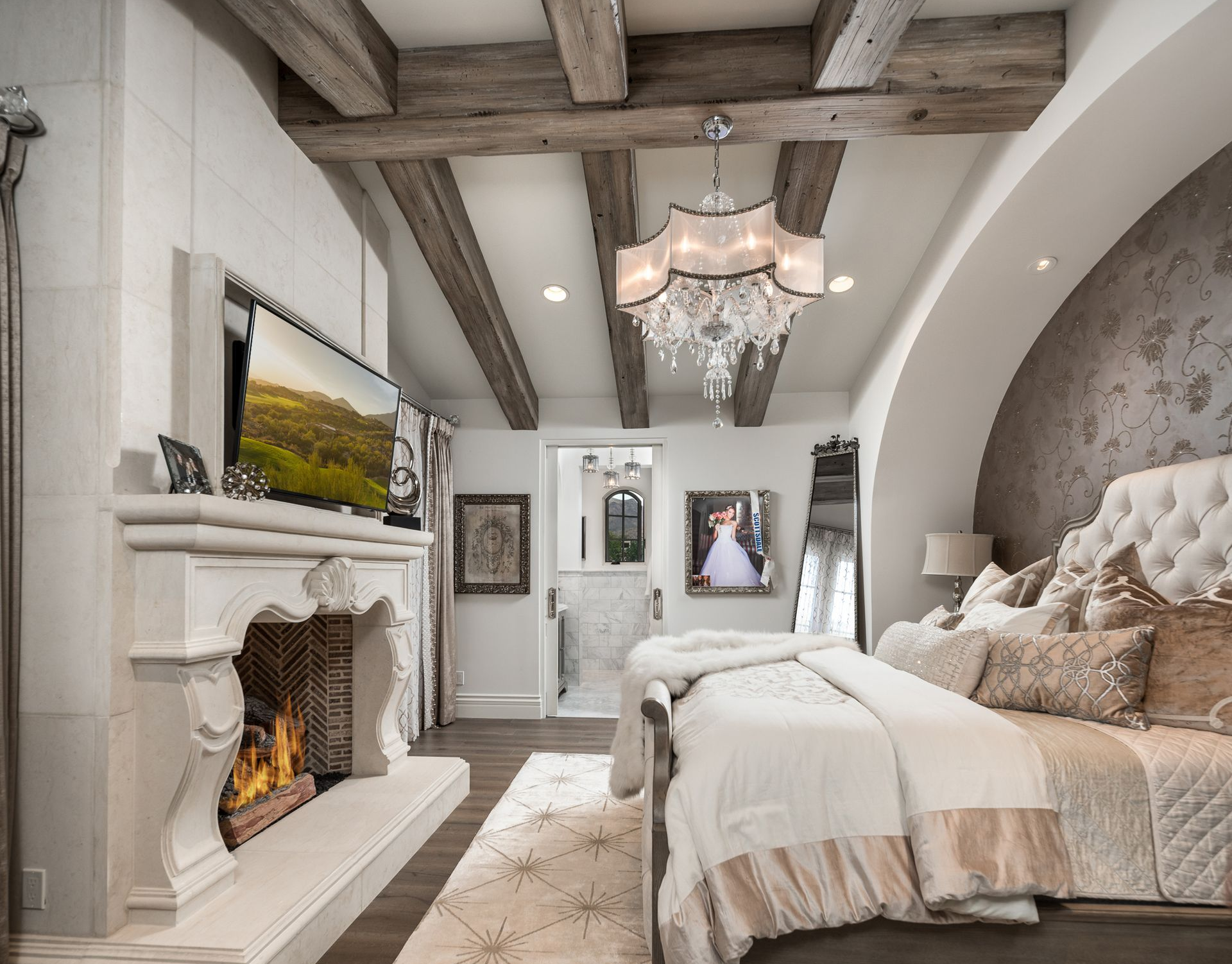 Elegant bedroom with fireplace, exposed wooden beams, chandelier, and a large tufted headboard.