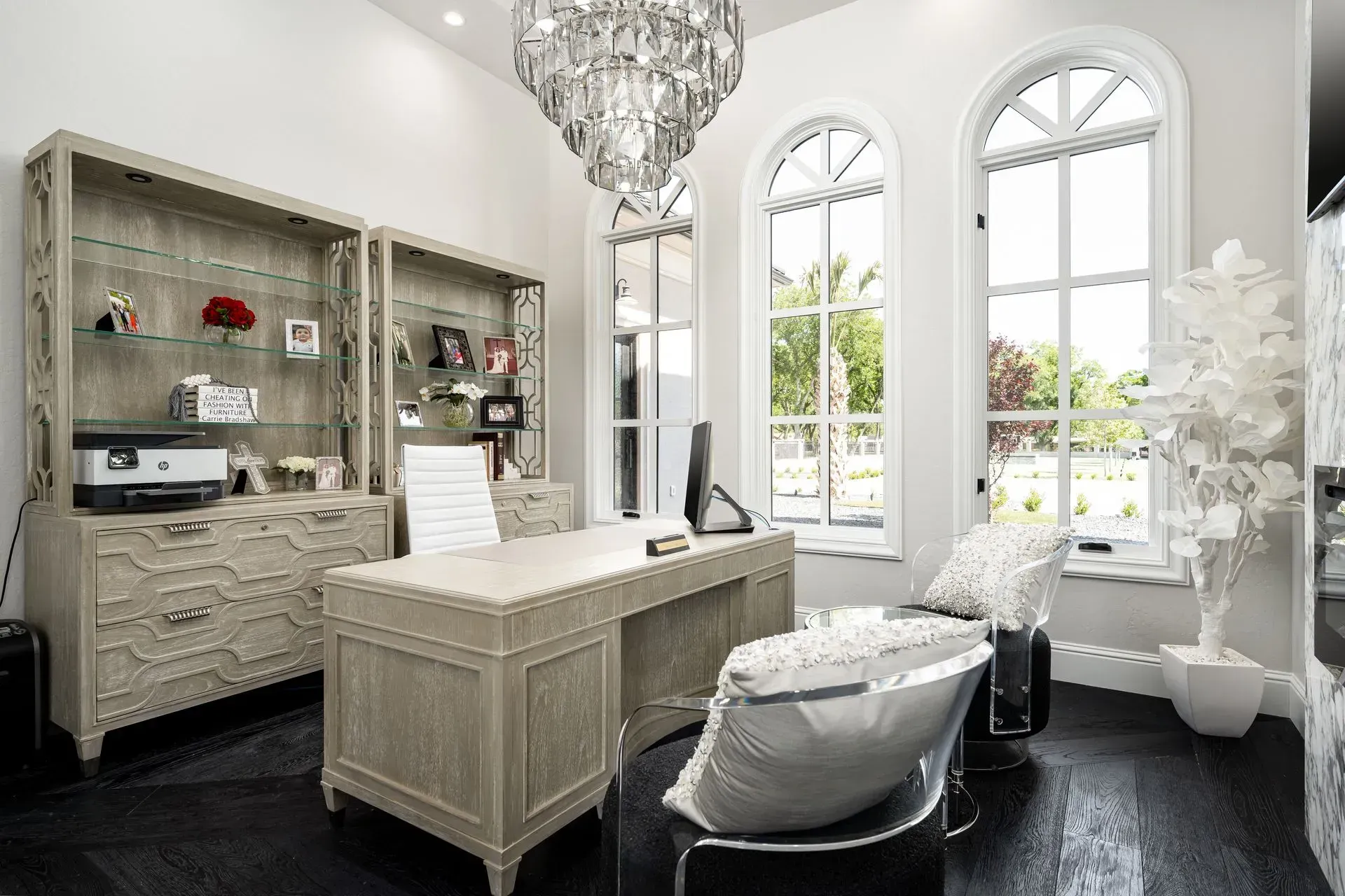 Elegant office with a desk, display cabinet, and large windows; black floor, white walls, and crystal chandelier.