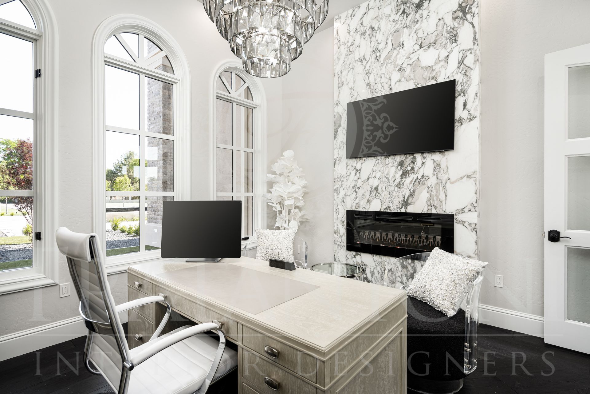 Elegant neutral home office with desk, marble fireplace, chandelier, and arched windows.