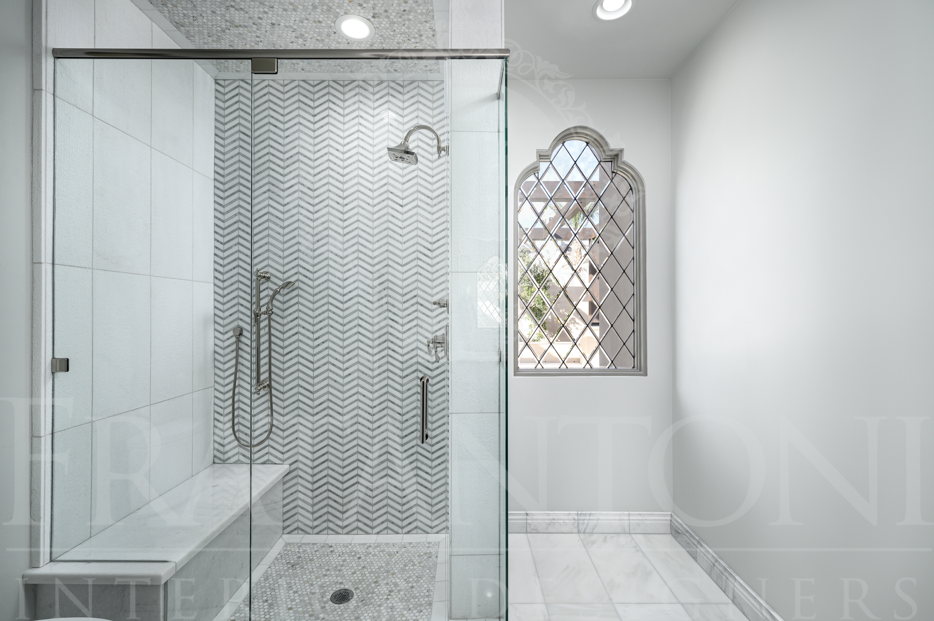 Modern luxury white bathroom with glass shower enclosure and decorative arched window. Featuring mosaic marble tiles, a shower bench, brass fixtures, and a custom window all designed by fratantoni interior designers