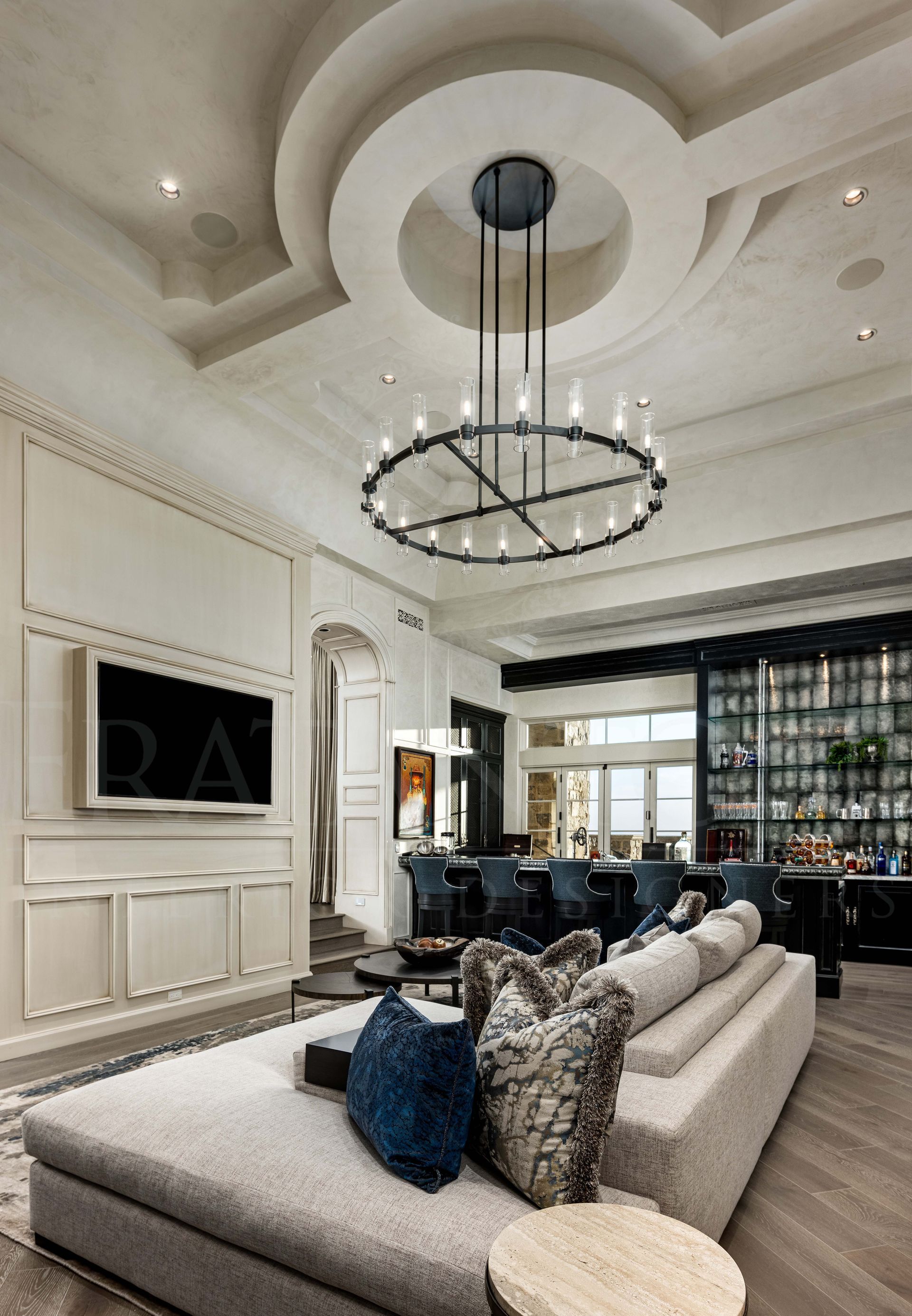 Luxury custom home bar with cream sofas, dark blue pillows, a circular chandelier, and a glass wall display.