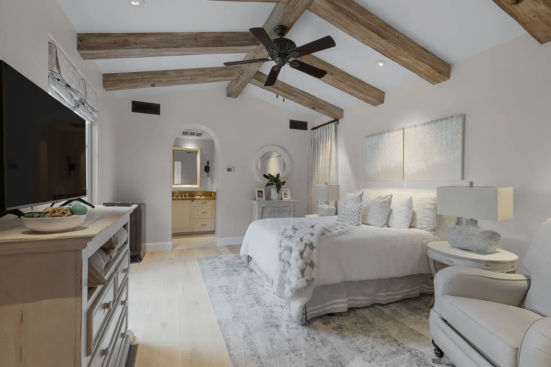 Bedroom with vaulted ceiling, exposed beams, neutral tones, bed, rug, and vanity visible.