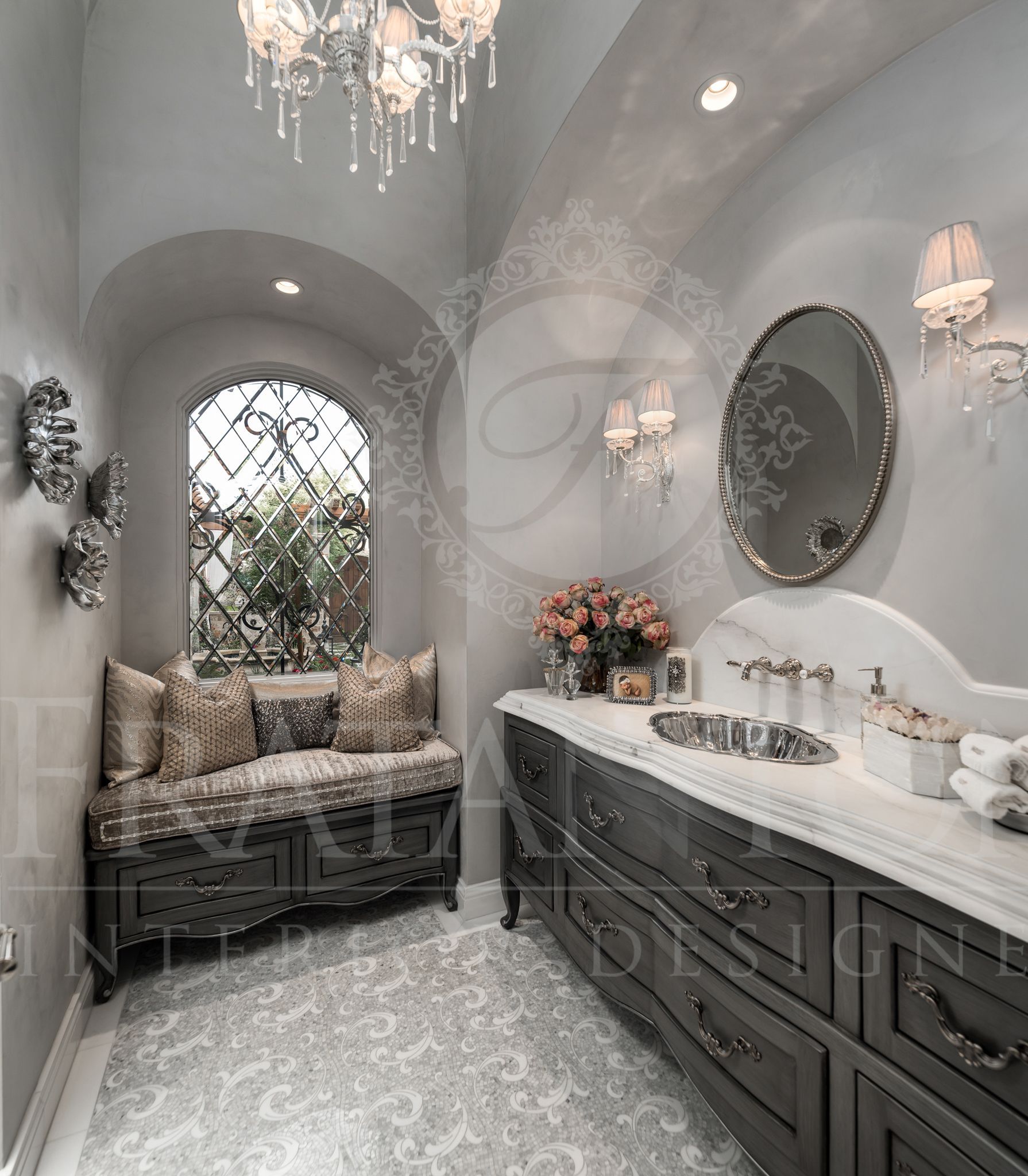 Elegant powder room with gray vanity, patterned mosaic floor tiles, arched window with seating, and chandelier.