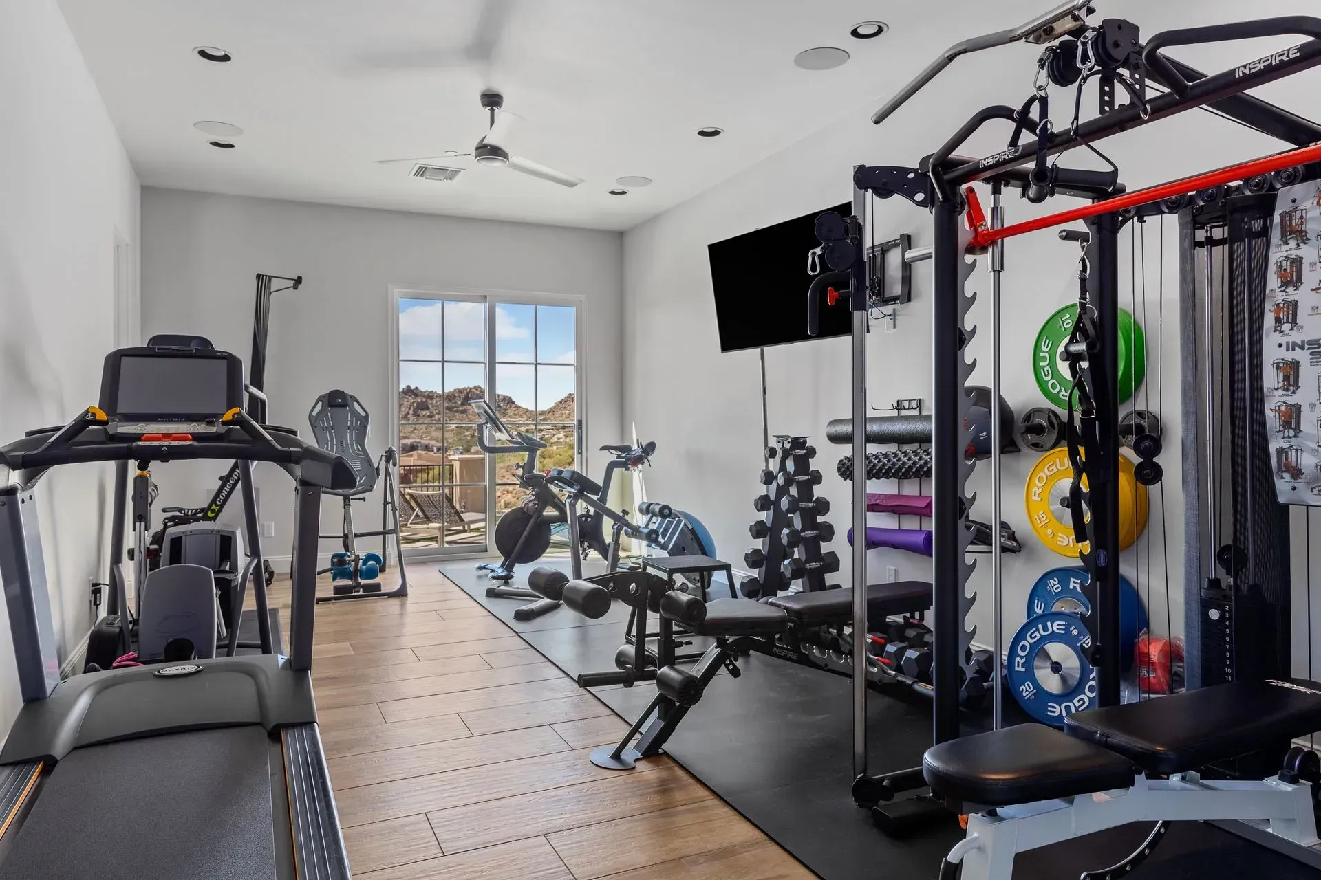 Home gym with exercise equipment: treadmill, weights, cable machine, bench, and window view.