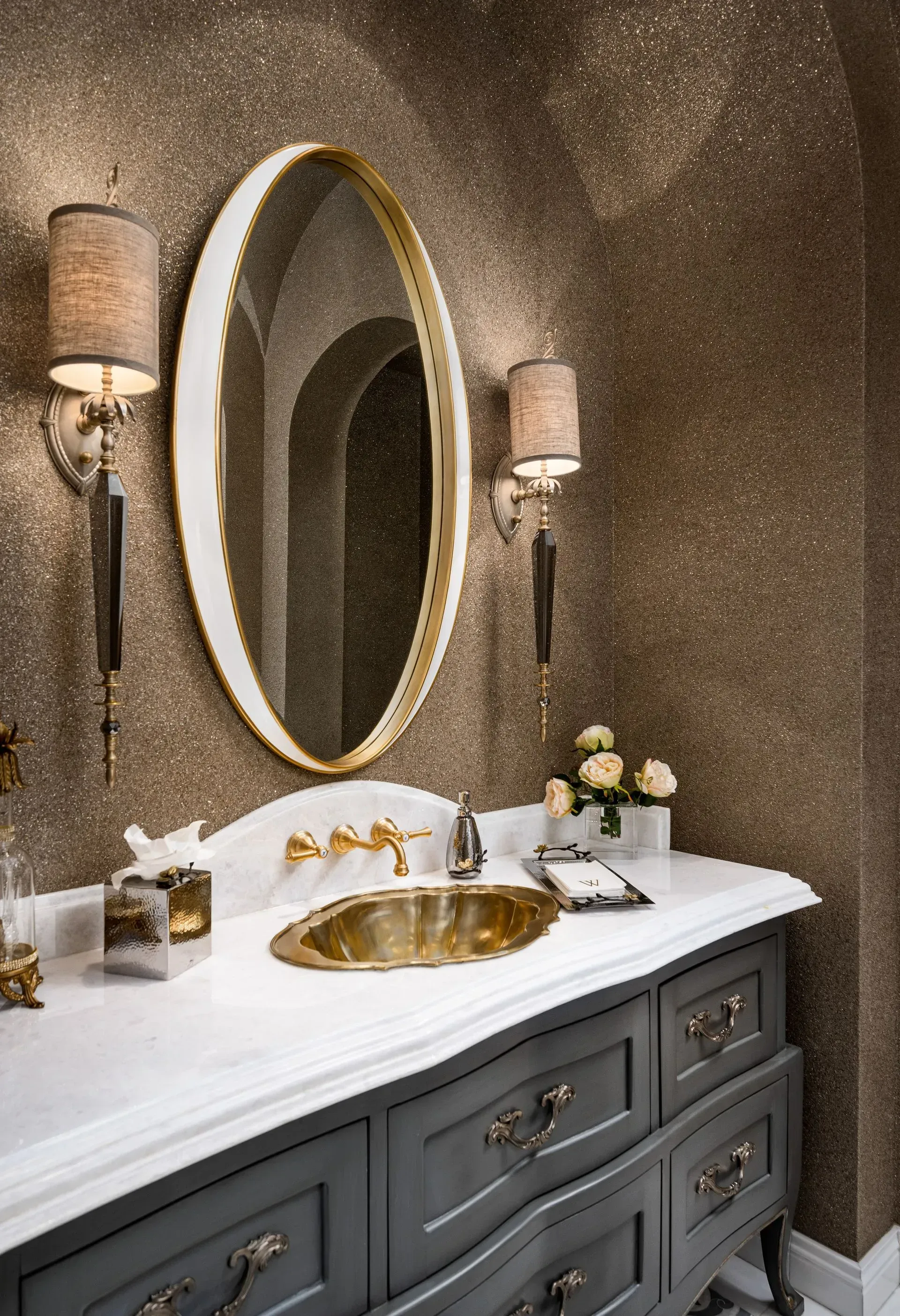 Elegant bathroom with gray vanity, gold fixtures, oval mirror, and shimmery wall.