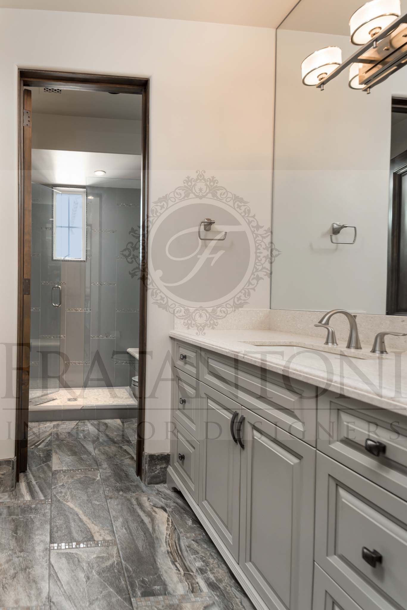 Bathroom vanity with marble countertop, gray cabinets, mirror, and overhead light fixtures with a separate toilet and shower room with shower bench