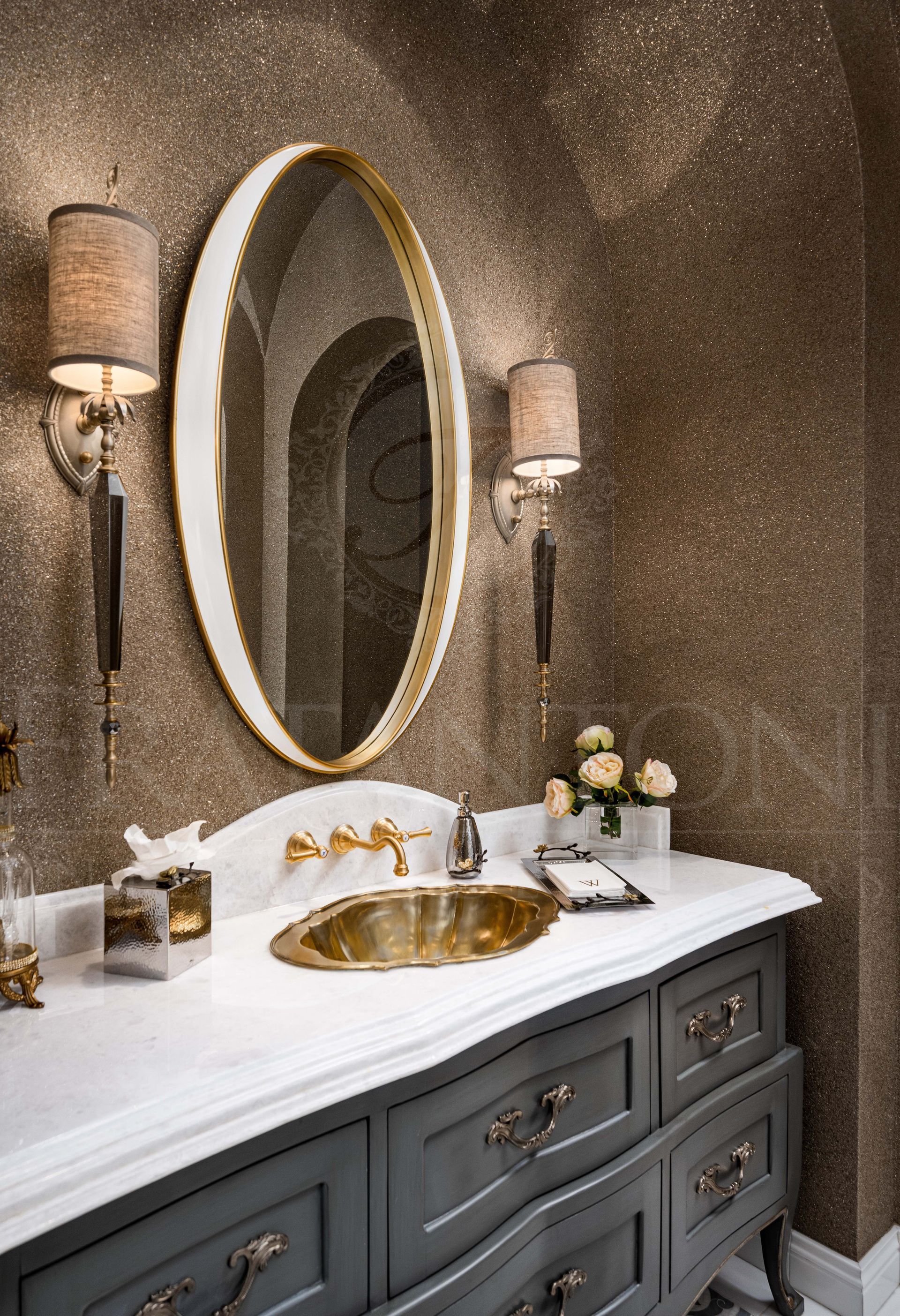 Custom bathroom vanity with oval mirror, gold faucet, gold drop-in sink, marble counter and backslash, ornate sconces, and glitter brown and gold walls