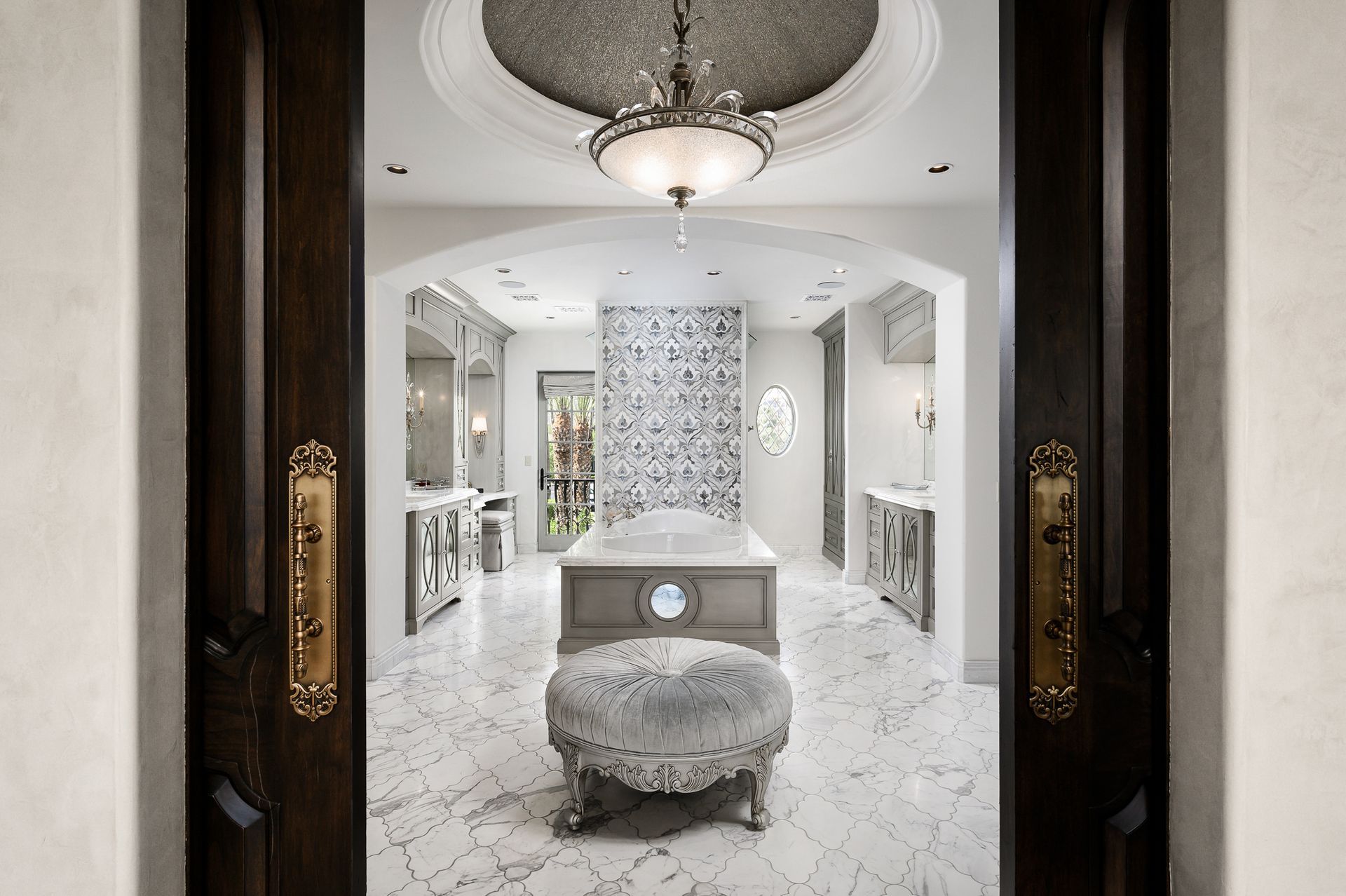 Elegant master bathroom, marble floors, dark wood doors, crystal chandelier, vanity, and ottoman.