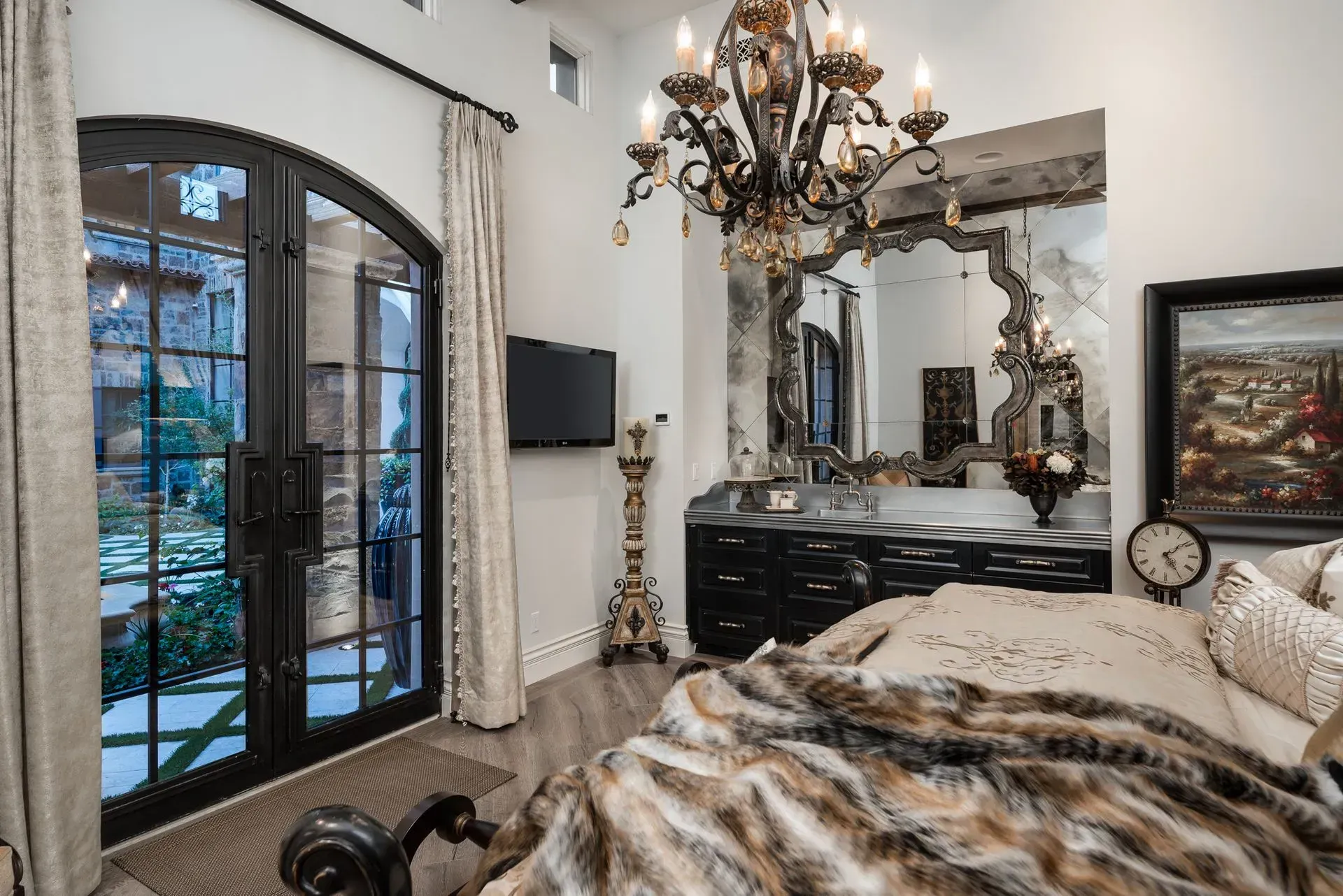 Bedroom with black door, ornate chandelier, large mirror, black dresser, and a furry blanket on the bed.
