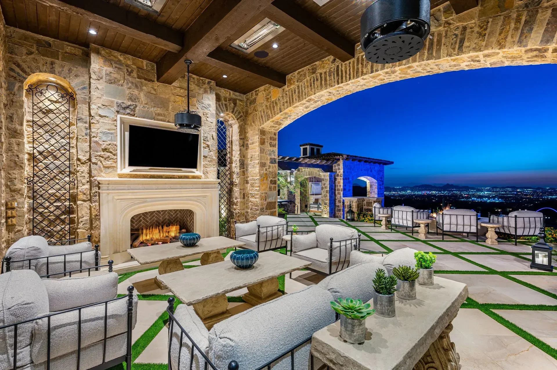 Outdoor patio with fireplace, TV, seating, and city view at dusk.