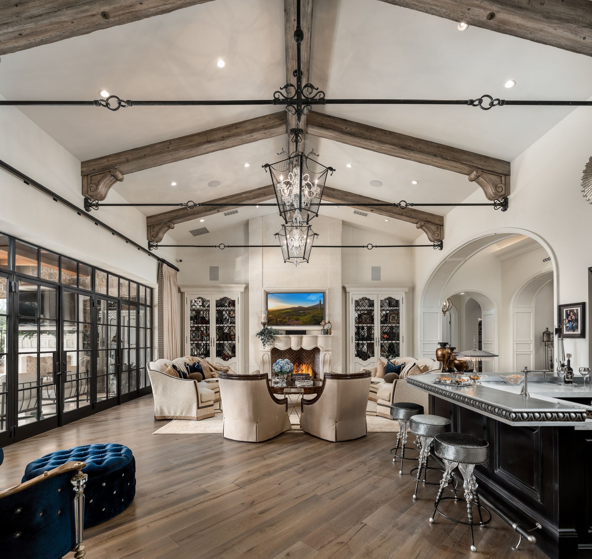 Elegant open-plan living room and kitchen with high vaulted beams, chandelier, fireplace, and bar seating