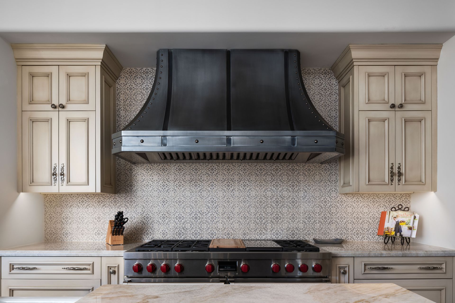Kitchen with range, metallic hood, cream cabinets, mosaic backsplash, and granite countertop.