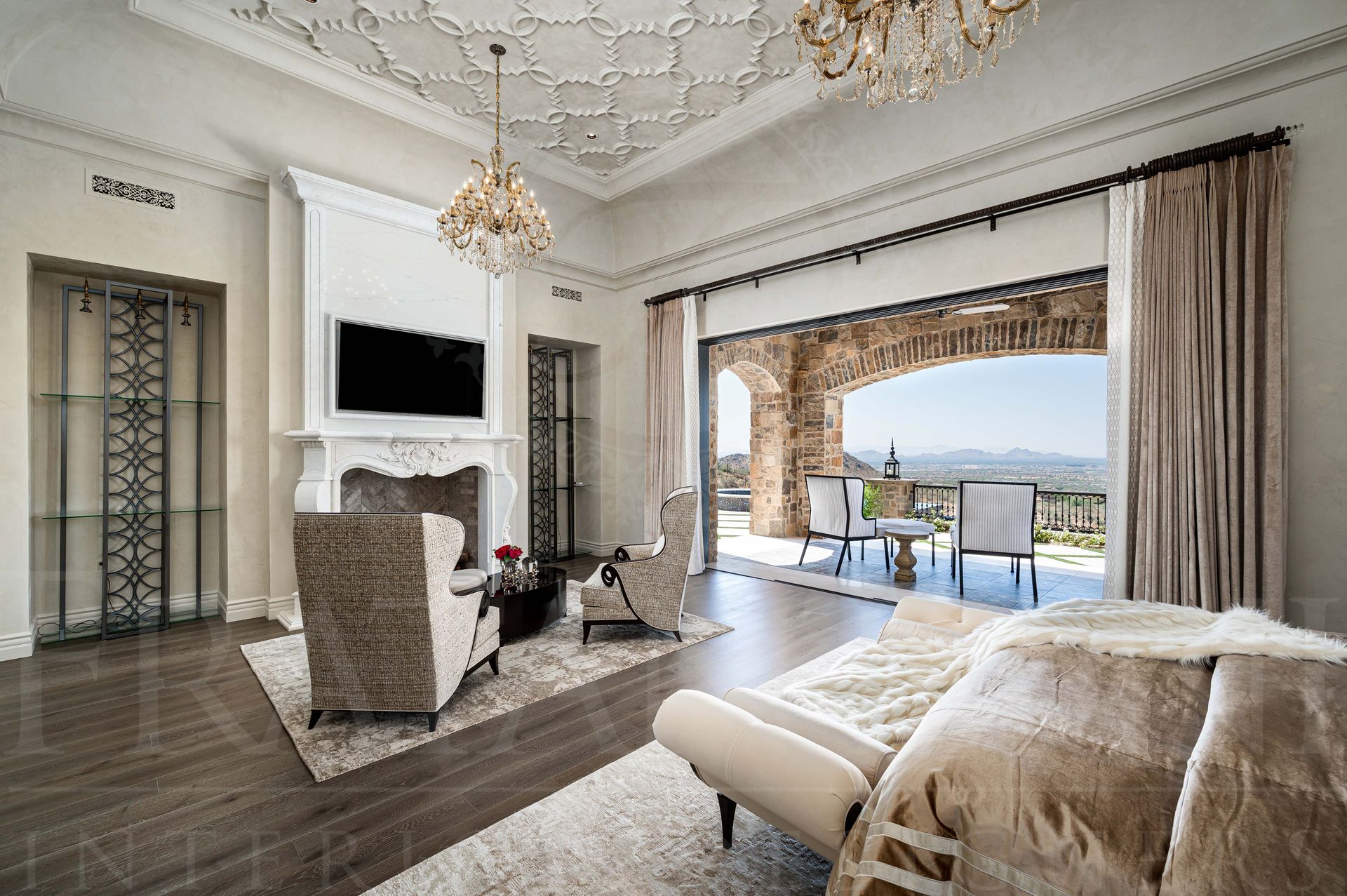 Luxury primary suite with chandeliers, carved-stone fireplace, TV, ornate ceiling details, plush chairs, and desert-view balcony doors