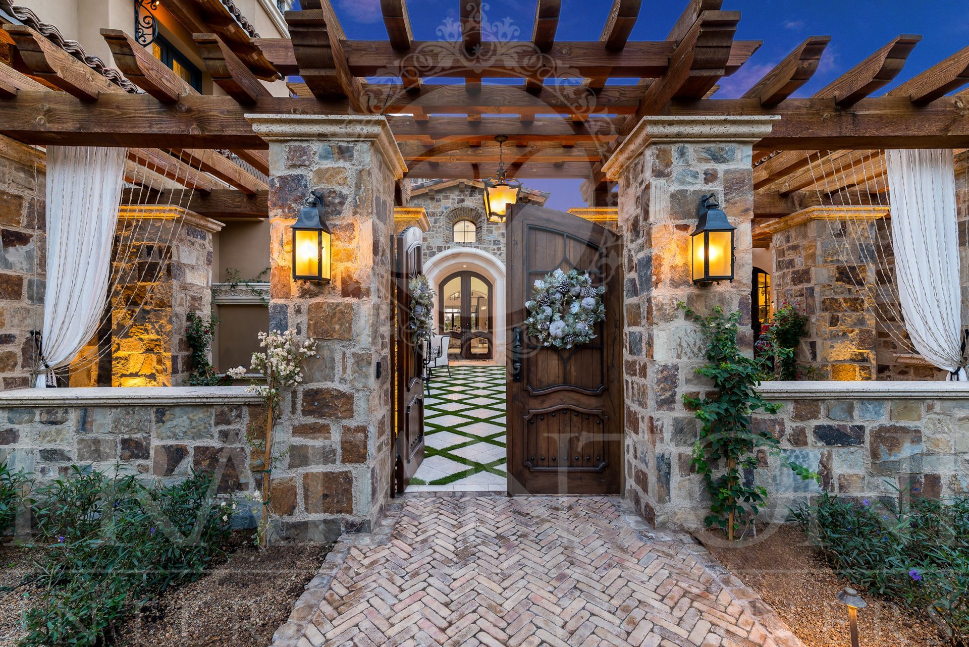 Stone facade entrance with open wooden door, decorated with a wreath, under a wooden pergola with outdoor curtains and entrance to a courtyard with geometric pavers and brick pavers designed by Fratantoni Interior Designers in scottsdale arizona.