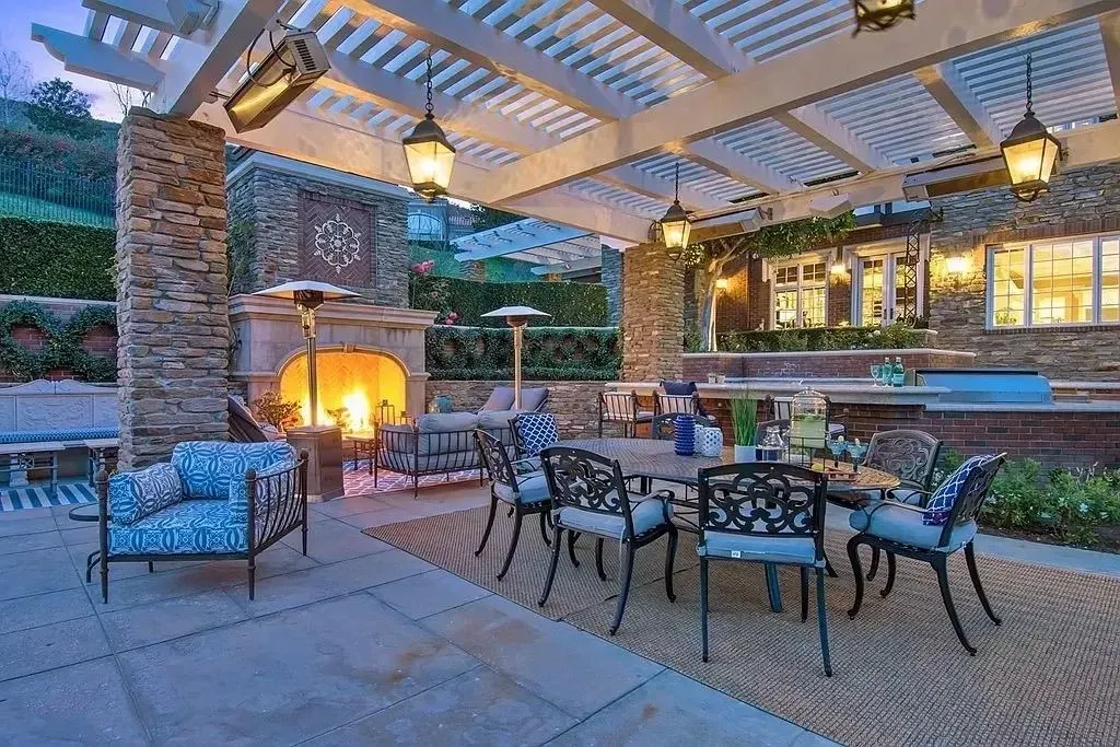 Outdoor patio with fireplace, dining table, and seating under a pergola. Evening setting, warm lighting.