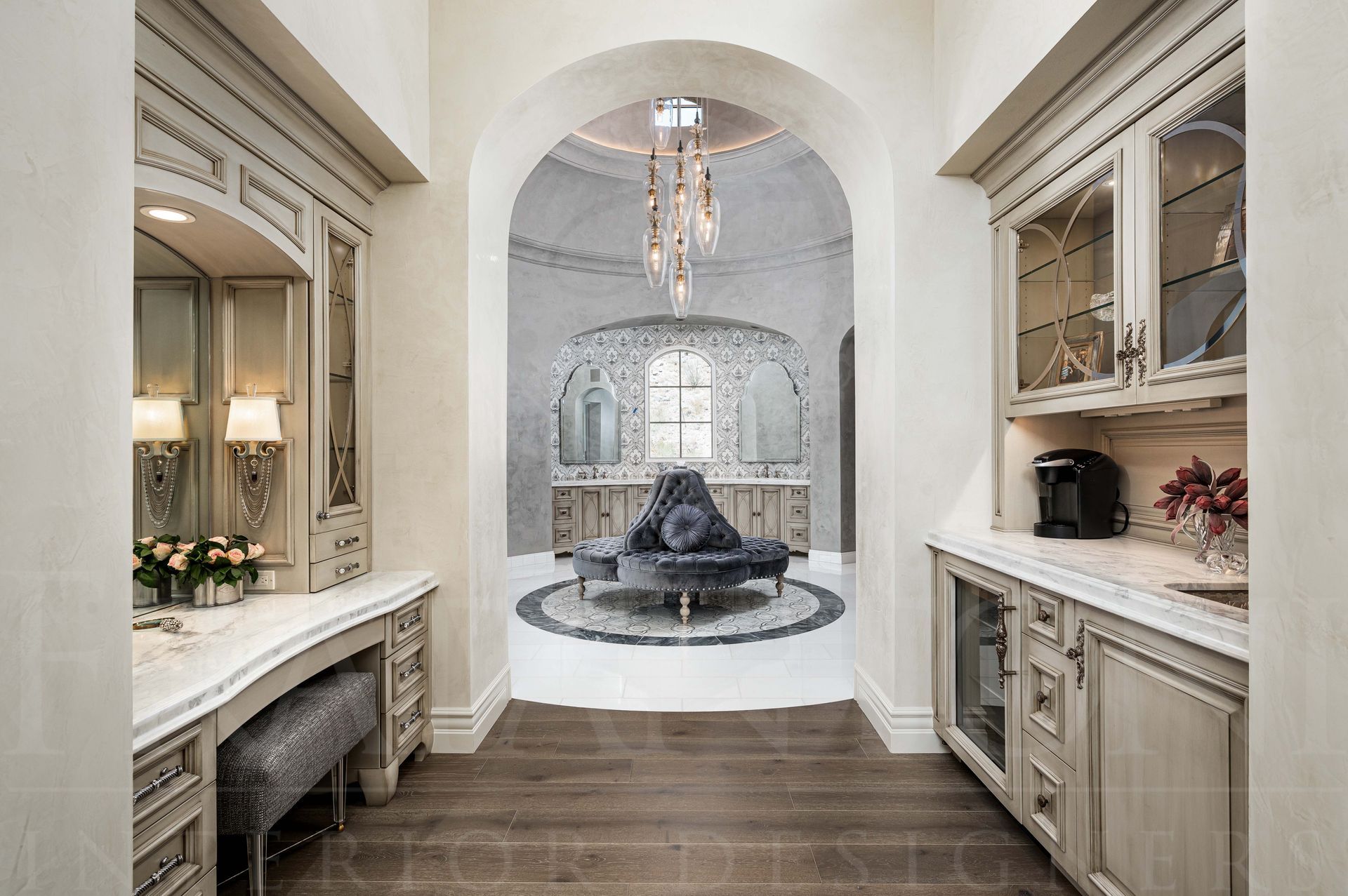 Elegant primary bath entry hallway with built-in cabinets, a mini kitchenette, a makeup vanity, leading to a chandelier-lit bathroom with custom seating and a domed ceiling