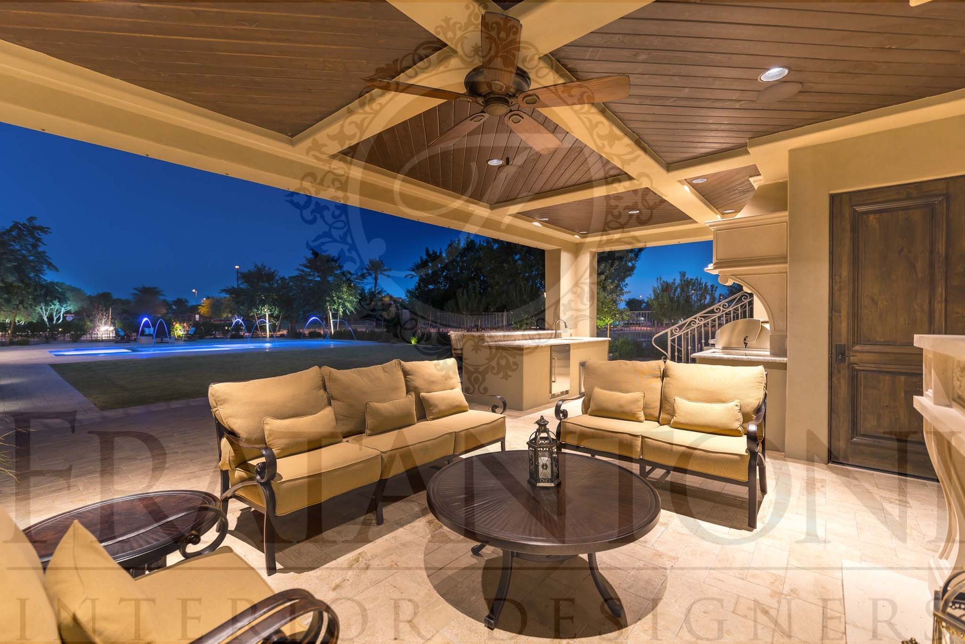Covered patio with custom seating, ceiling fan, coffered ceilings, and round wooden table overlooking a lit pool at dusk