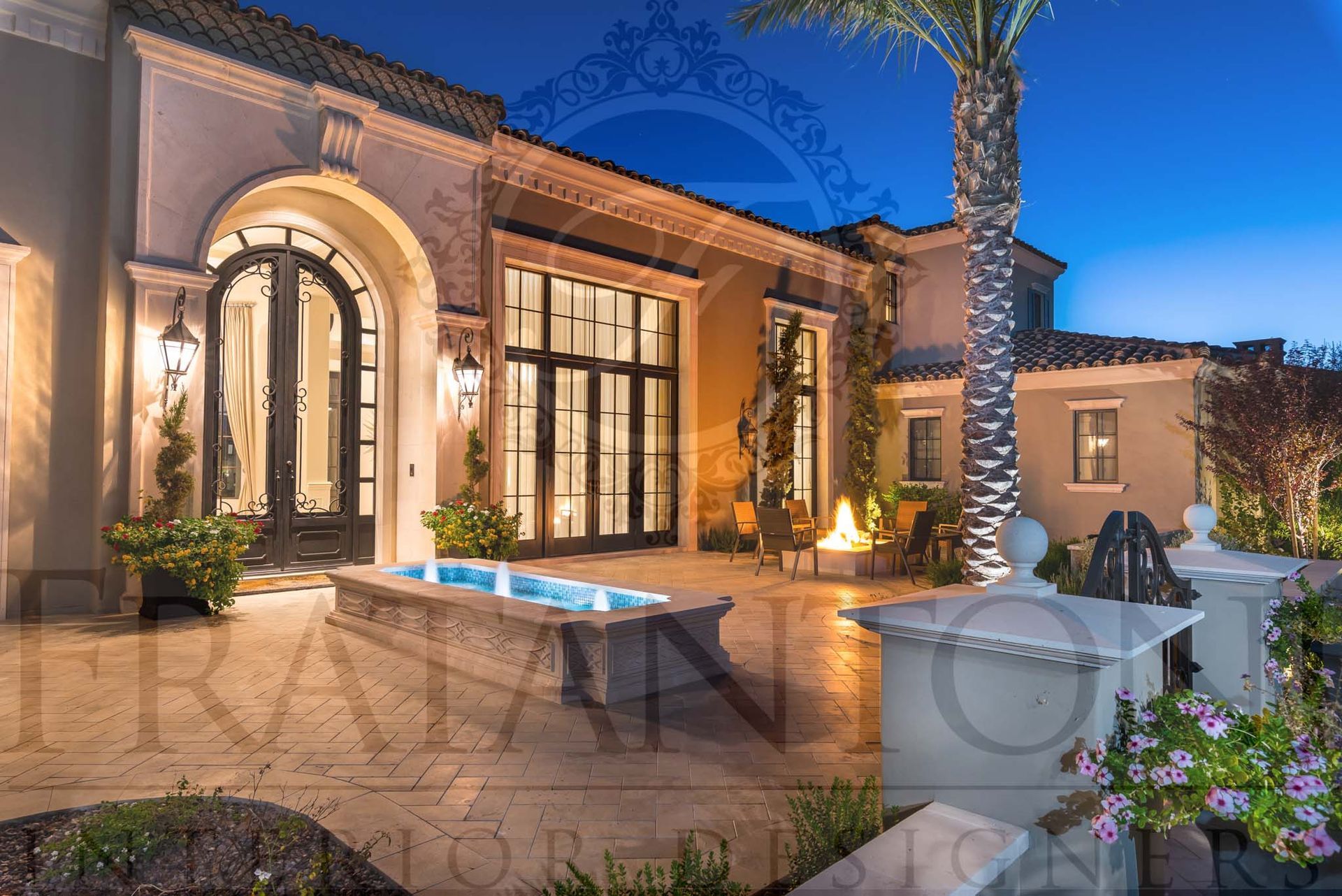 Luxury villa courtyard at dusk with arched doors, large windows, exterior lighting, a fire pit, natural stone pavers, lit fountain, and palm trees in paradise valley arizona designed by fratantoni interior designers