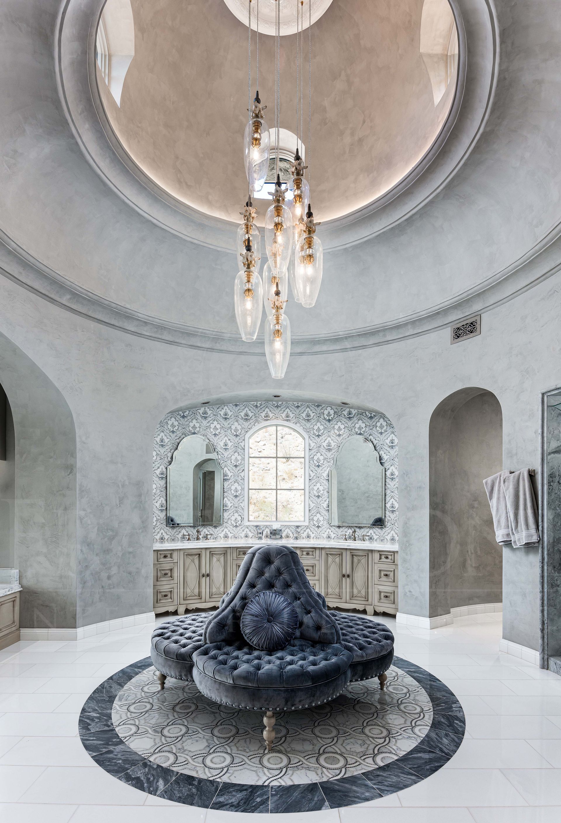 Elegant custom primary bathroom with a dramatic domed ceiling, with ornate chandelier, arched alcoves, and dark sculptural sofa on a round inlaid mosaic floor designed by Fratantoni Interior Designers