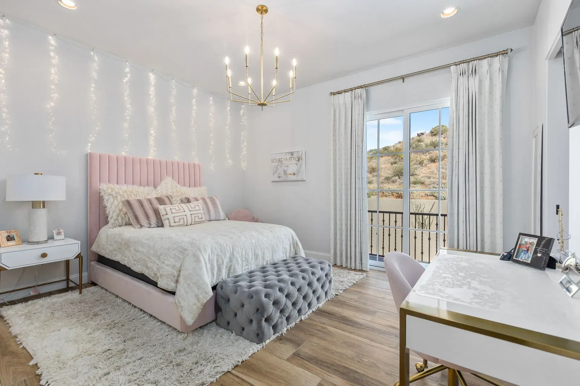 A pink and white bedroom with a bed, desk, and mountain view. String lights and a chandelier hang from the ceiling.