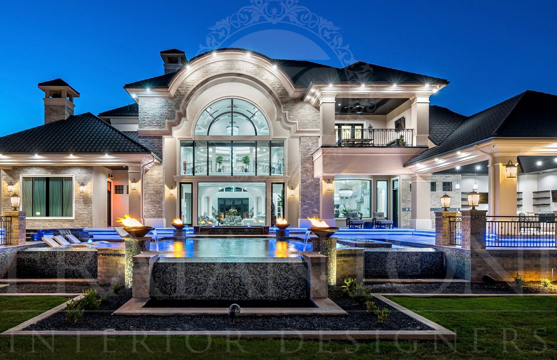 Luxury mansion exterior at dusk, illuminated pool, stone facade, exterior lighting, an infinity edge pool, and outdoor fireplaces.