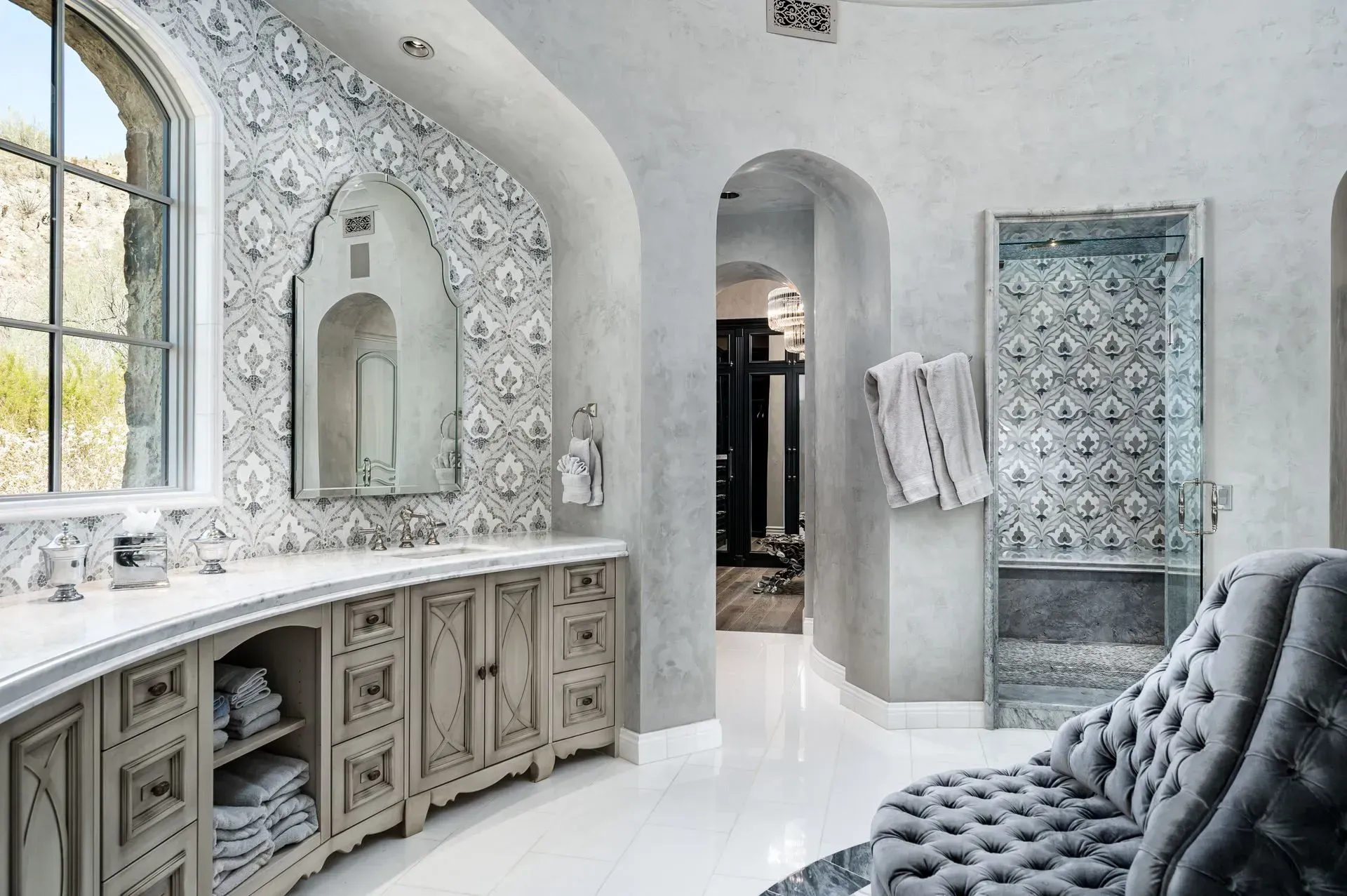 Elegant bathroom with patterned tile, arched mirror, curved vanity, and tufted gray chair.