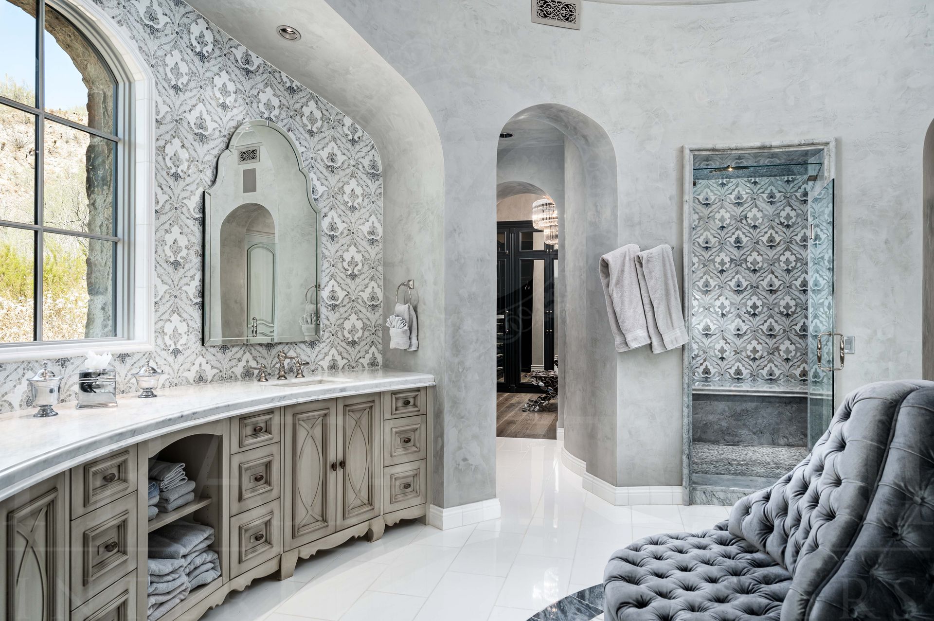 Bright custom master bathroom with mosaic tile, arched mirrors, arched doorways, a large custom shower, vanity cabinets, and a tufted chair.