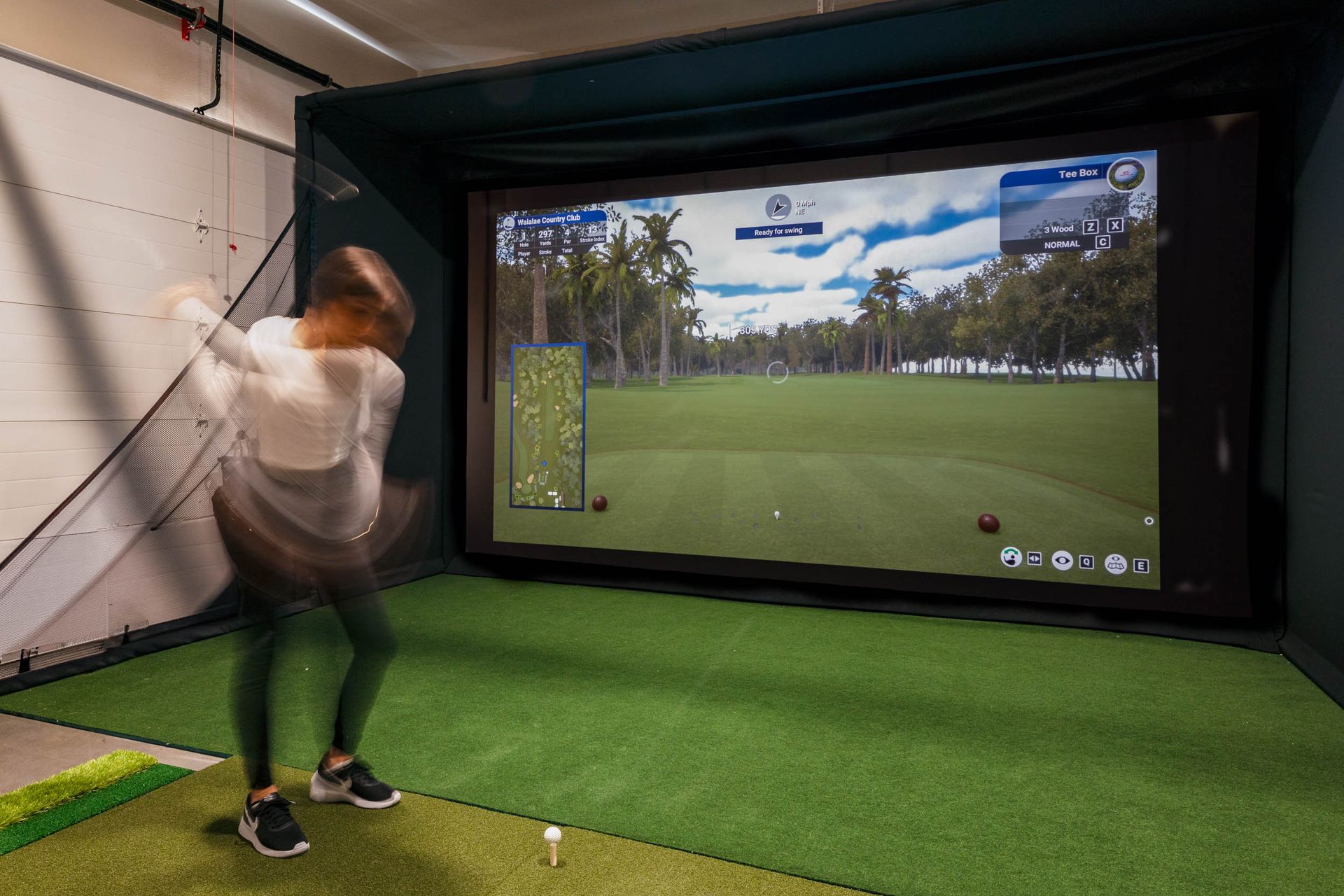 Woman swinging a golf club in a golf simulator room, with a projected golf course on a large screen.
