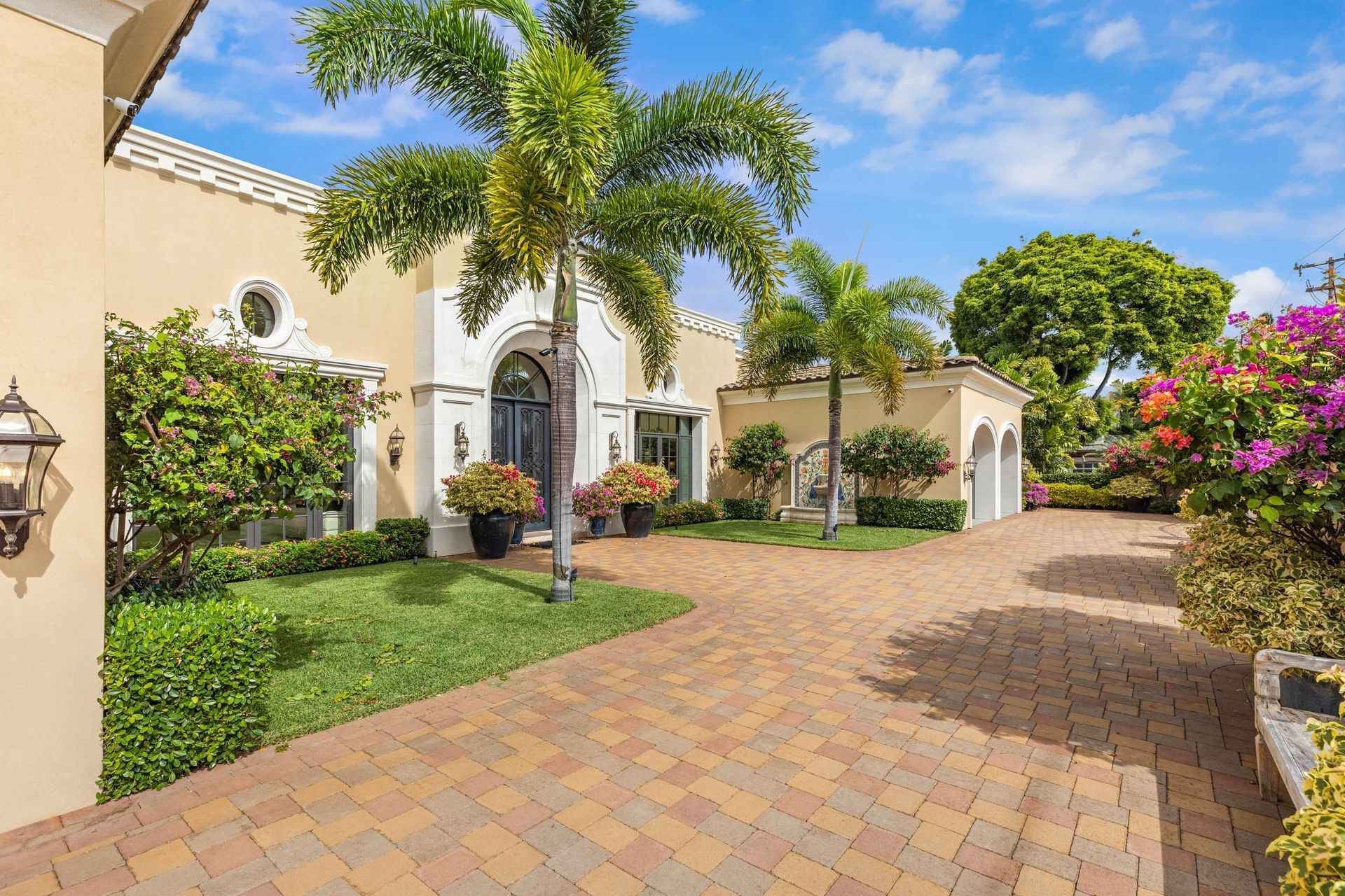 Beige stucco home with brick driveway and lush landscaping under a blue sky.