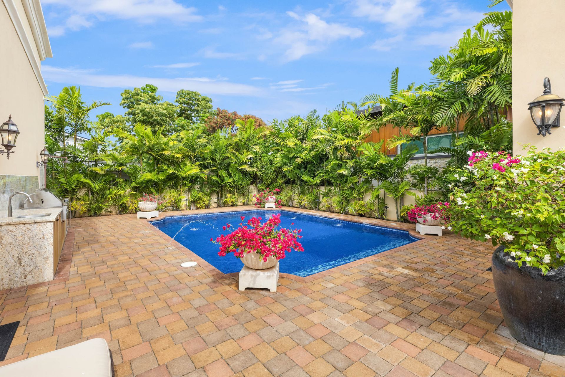 Backyard with a blue pool, surrounded by green foliage, brick patio, and outdoor kitchen.