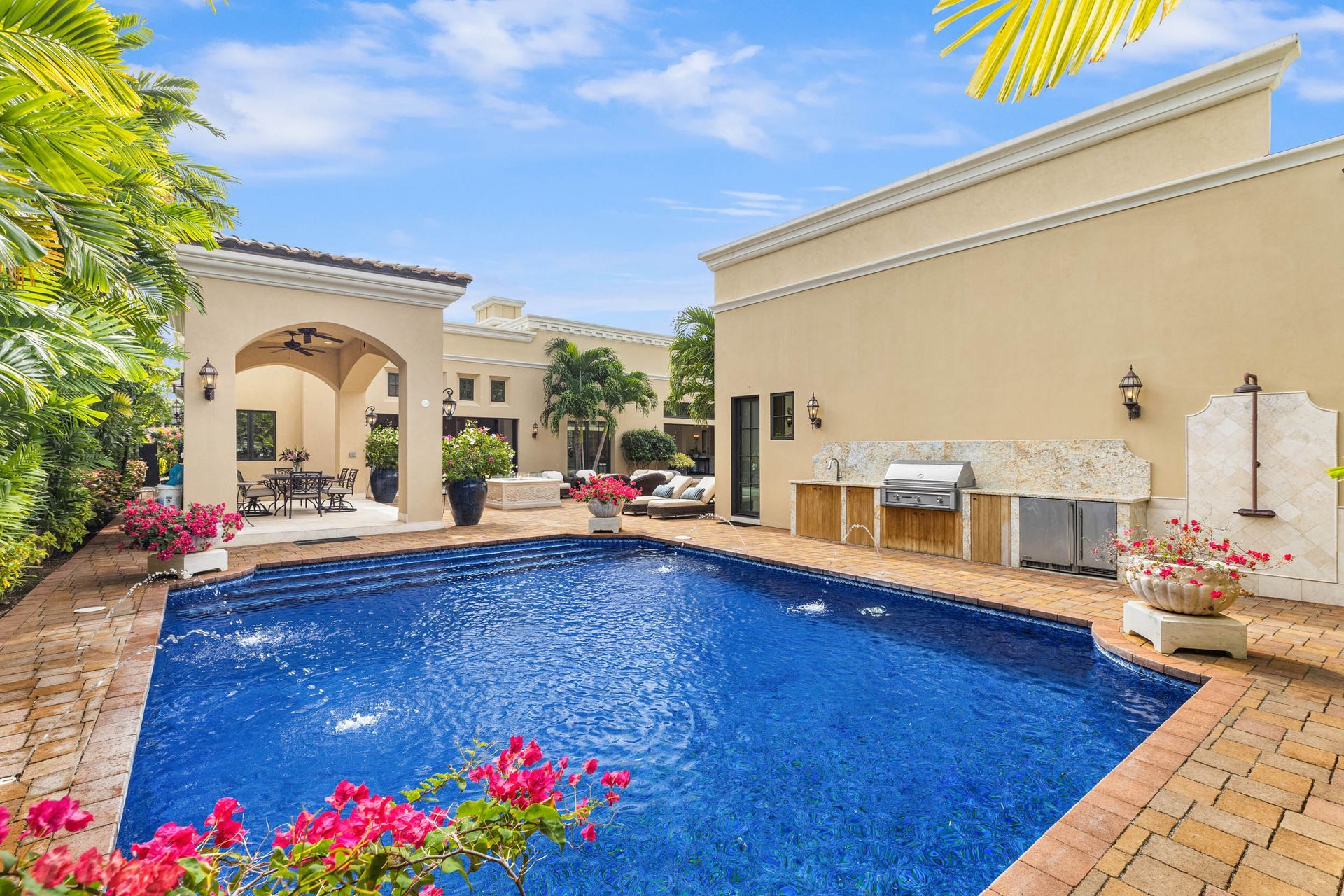 A backyard pool with a covered patio, outdoor kitchen, and landscaping under a blue sky.