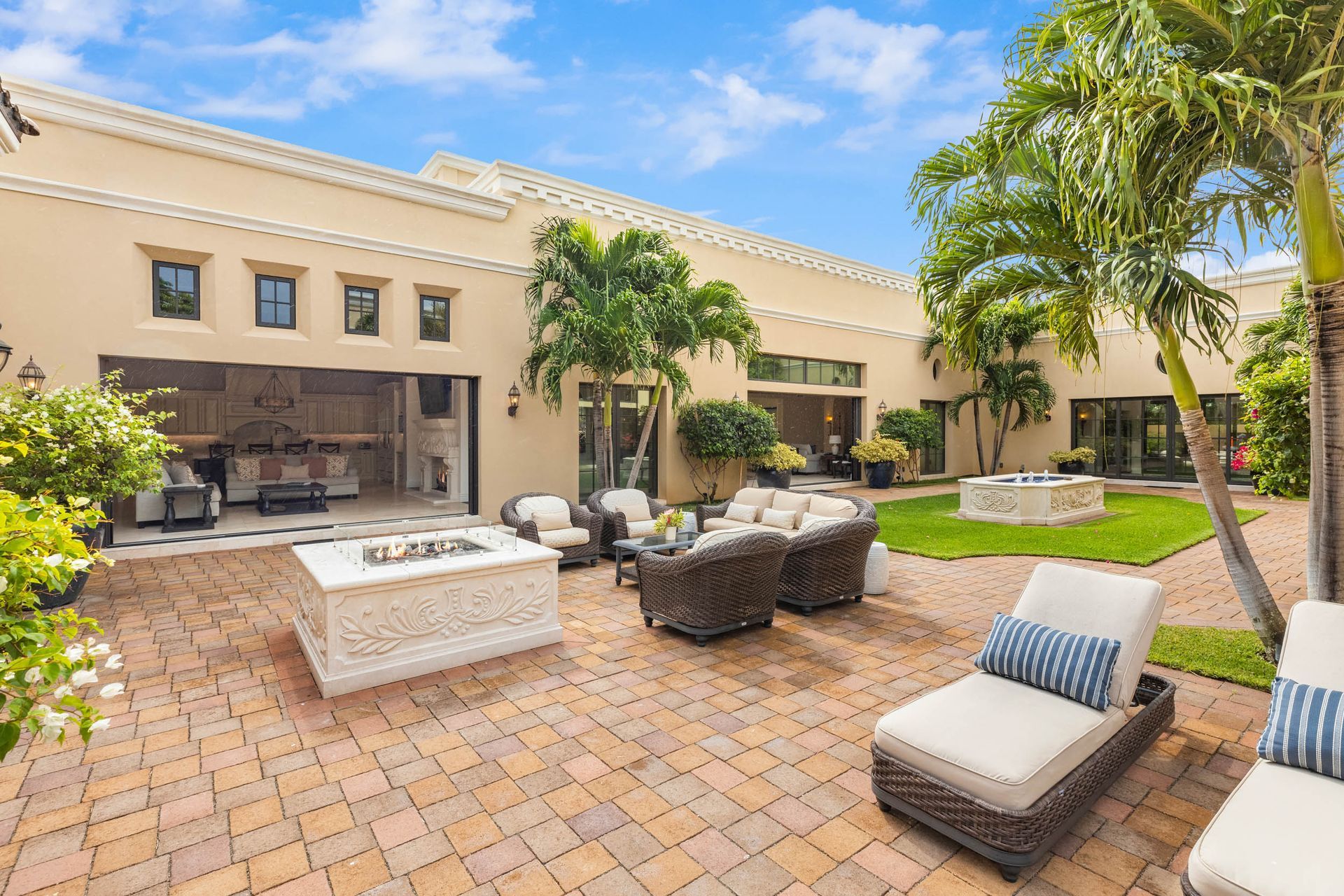 Patio with outdoor seating, brick pavers, and fire pit in a courtyard, flanked by a stucco house and palm trees.
