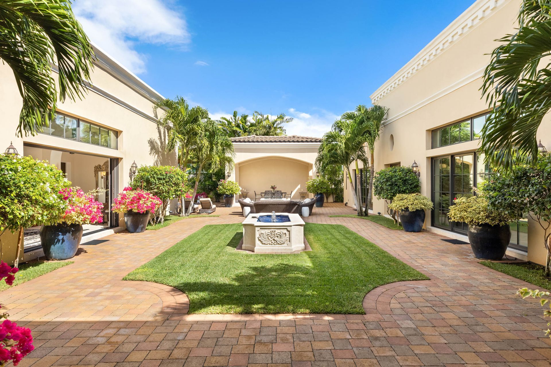 Courtyard with brick patio, green grass, and potted plants, surrounded by cream-colored buildings under a blue sky.