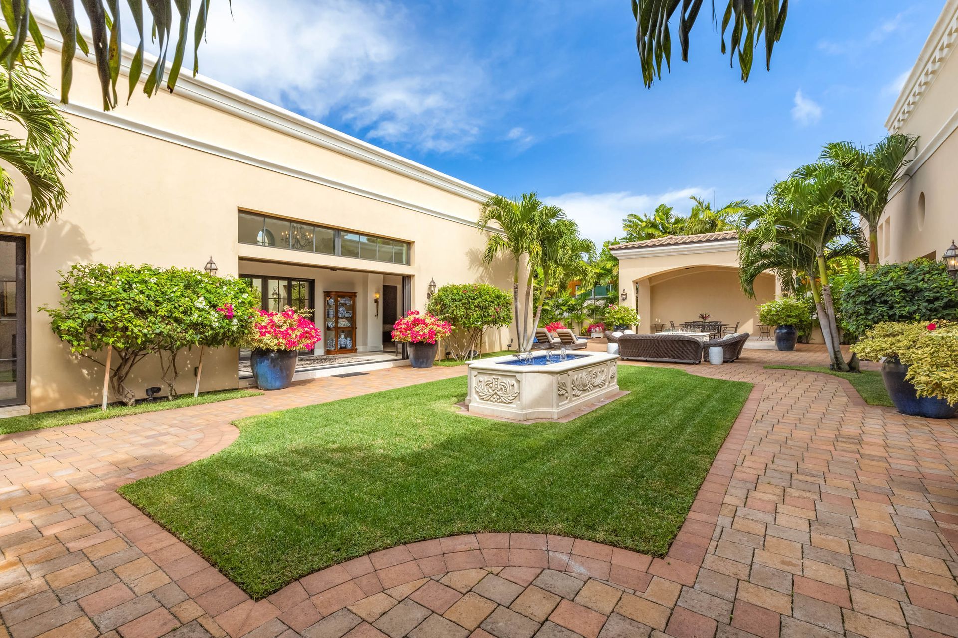 Courtyard with green lawn, fountain, and brick pathway surrounded by cream-colored buildings and palm trees.