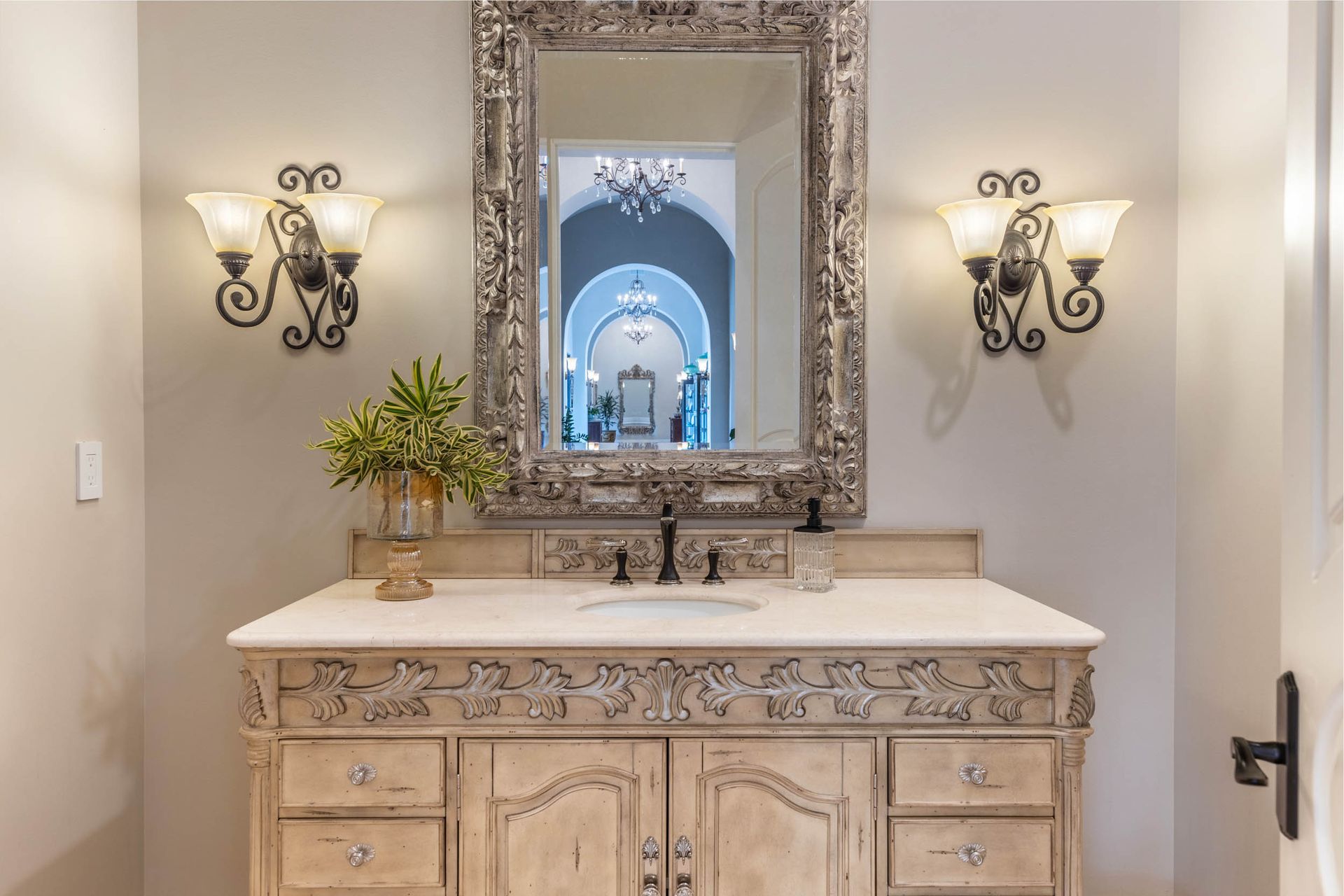 Ornate vanity with mirror, sconces, and floral arrangement in a neutral-toned bathroom.