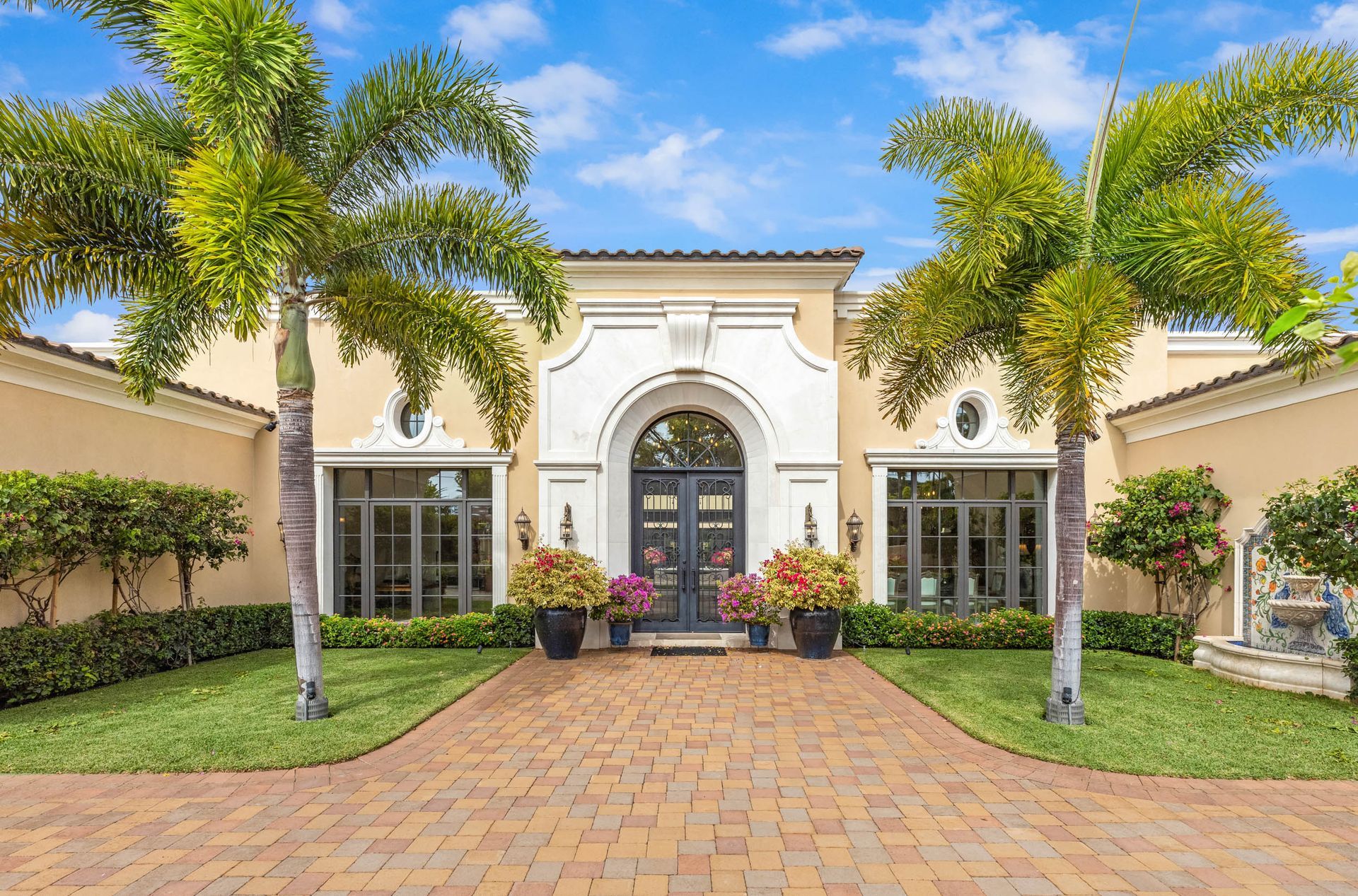 Elegant beige home with brick driveway, arched doorway, and palm trees.