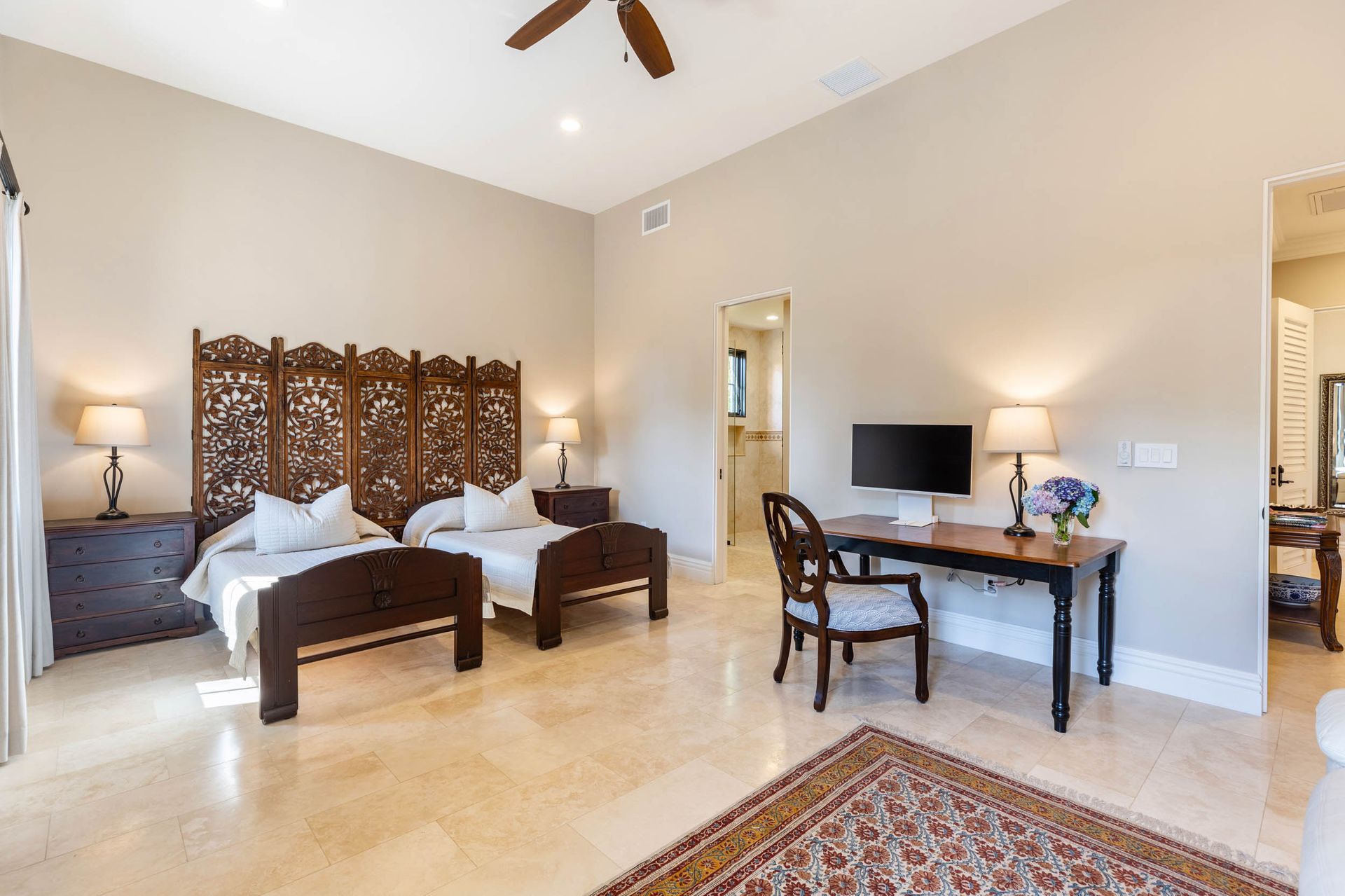 Spacious bedroom with two beds, wooden headboard screen, desk, lamps, and beige walls and flooring.