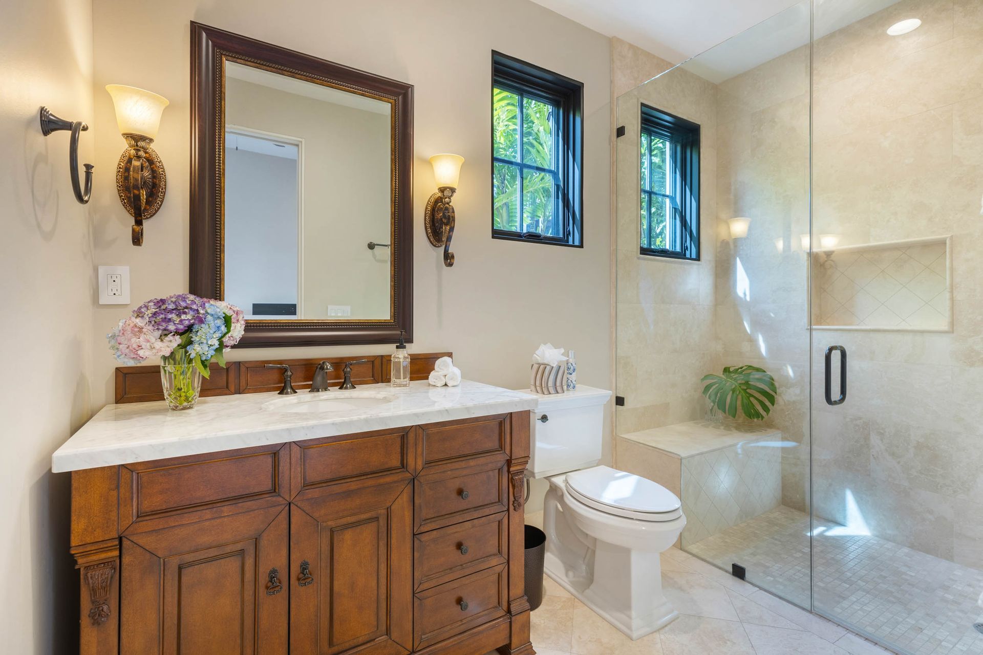 Bathroom with wood vanity, marble countertop, large mirror, glass shower, toilet, and two wall sconces.