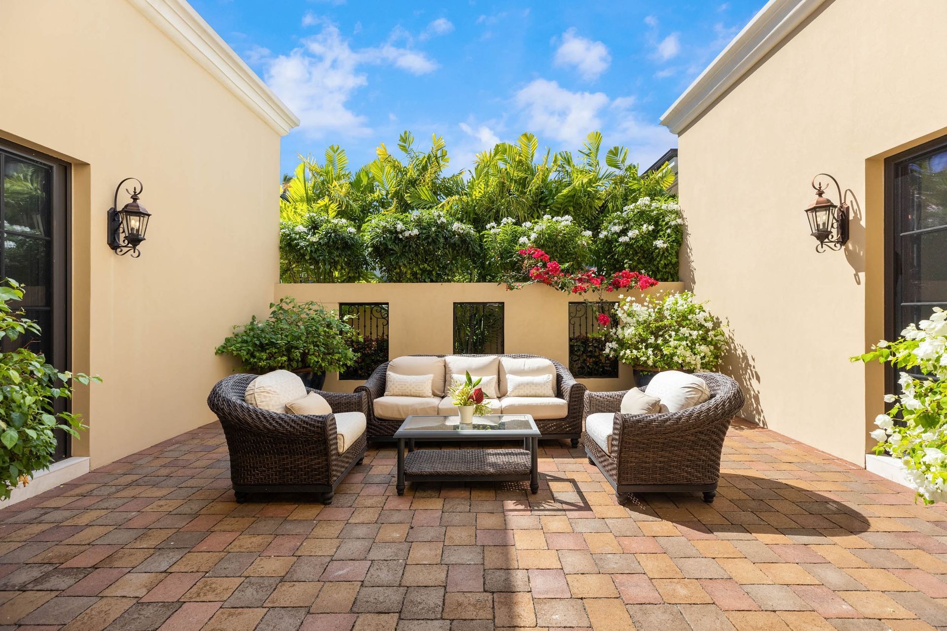 Patio with wicker furniture, brick floor, surrounded by walls and greenery under a blue sky.