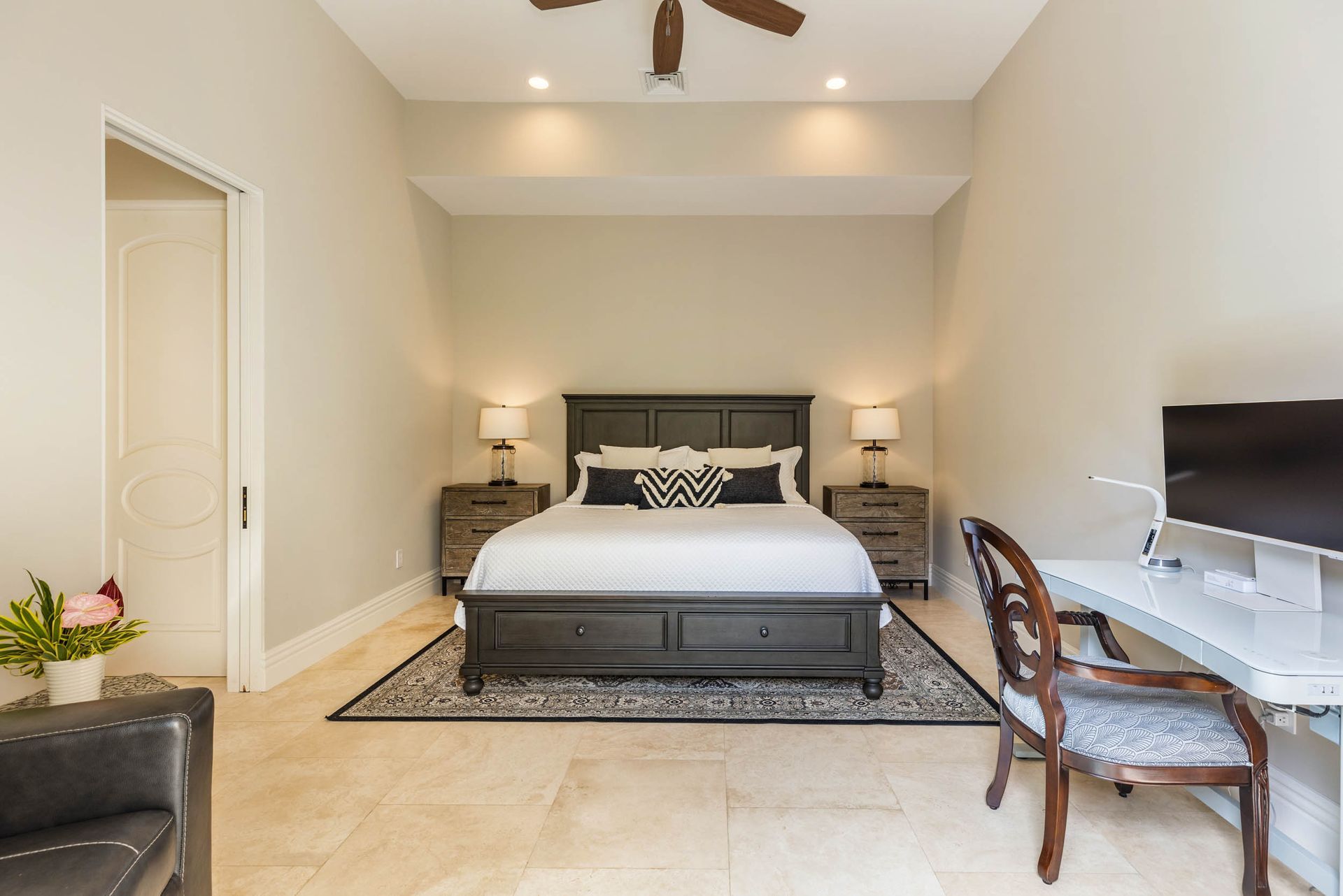 Bedroom with bed, nightstands, desk, and chair. Beige walls, stone floor, and decorative pillows.